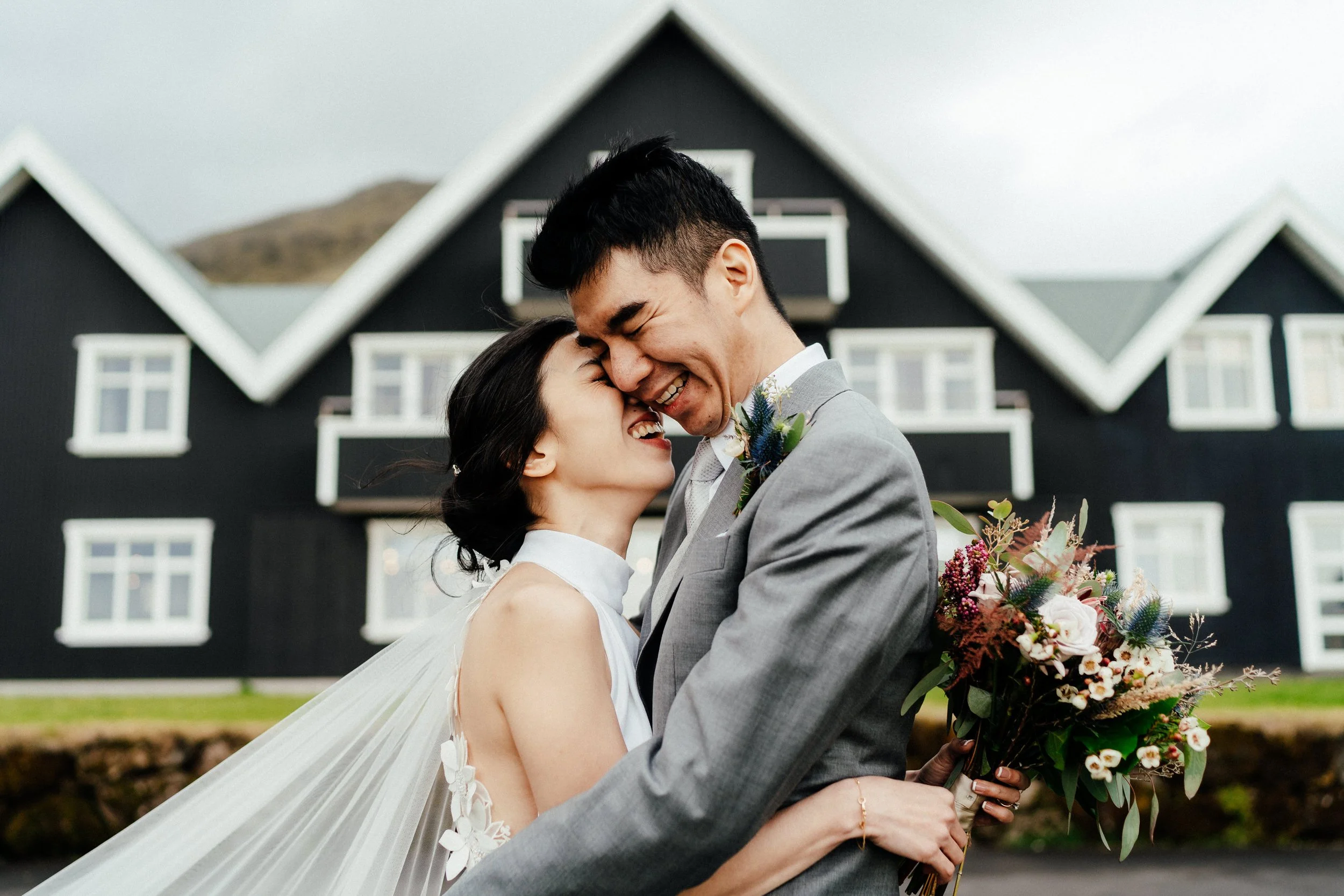 Newlywed couple embracing and smiling outside a black-and-white house, with the bride holding a colorful bouquet.