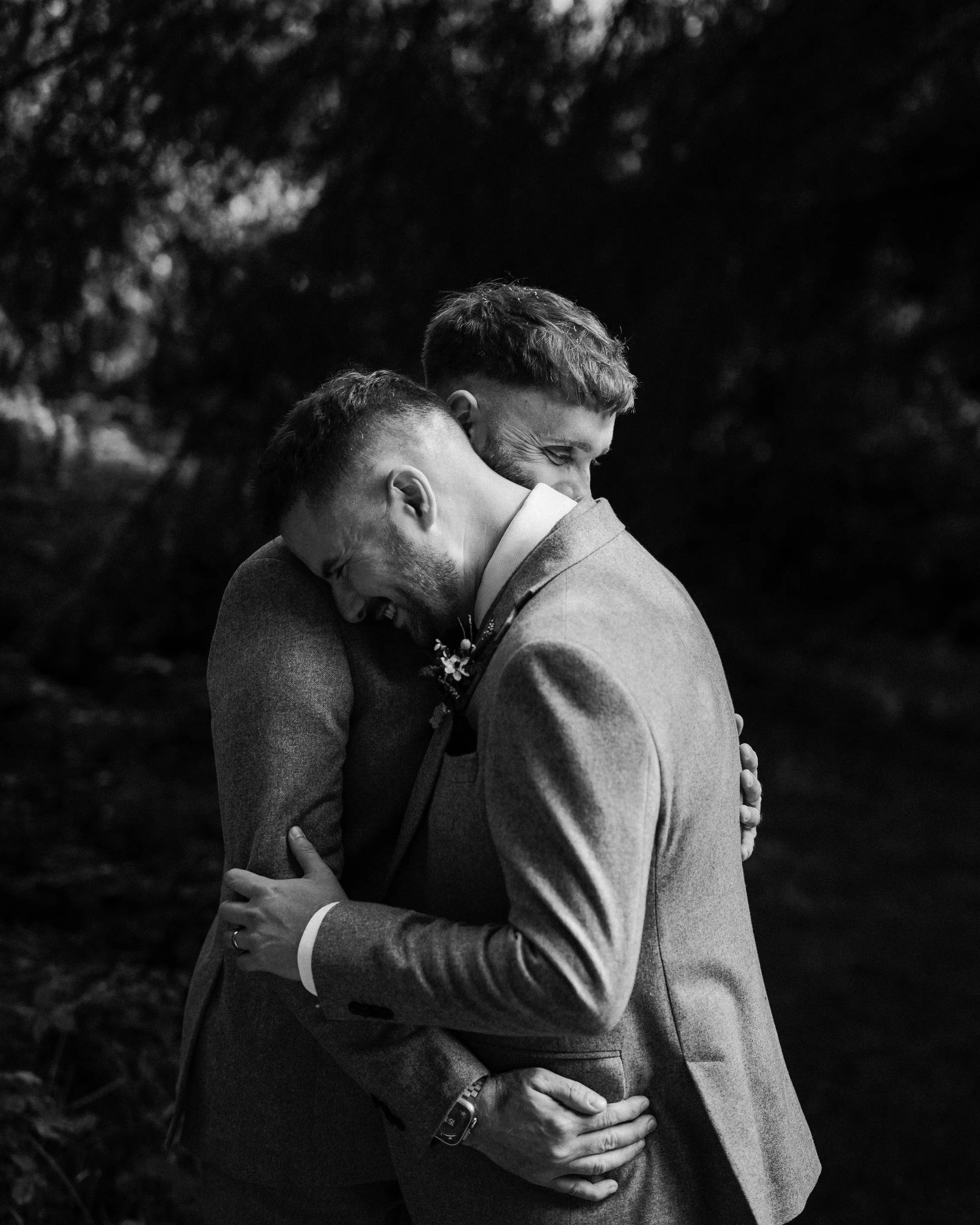 Black-and-white wedding portrait of two men embracing outdoors, wearing suits.