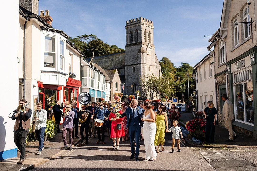 Wedding procession with a marching band walking through a village street past a historic stone church in England