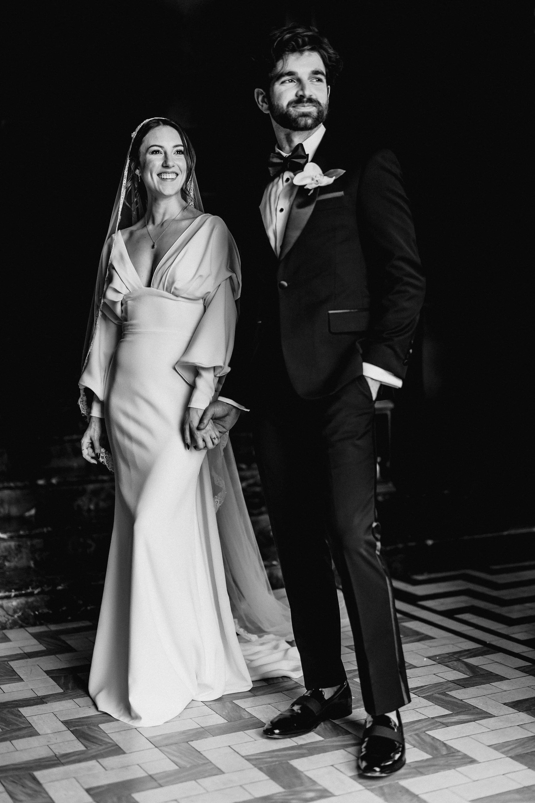Black-and-white wedding portrait of a smiling bride and groom holding hands, wearing a wedding gown and tuxedo.