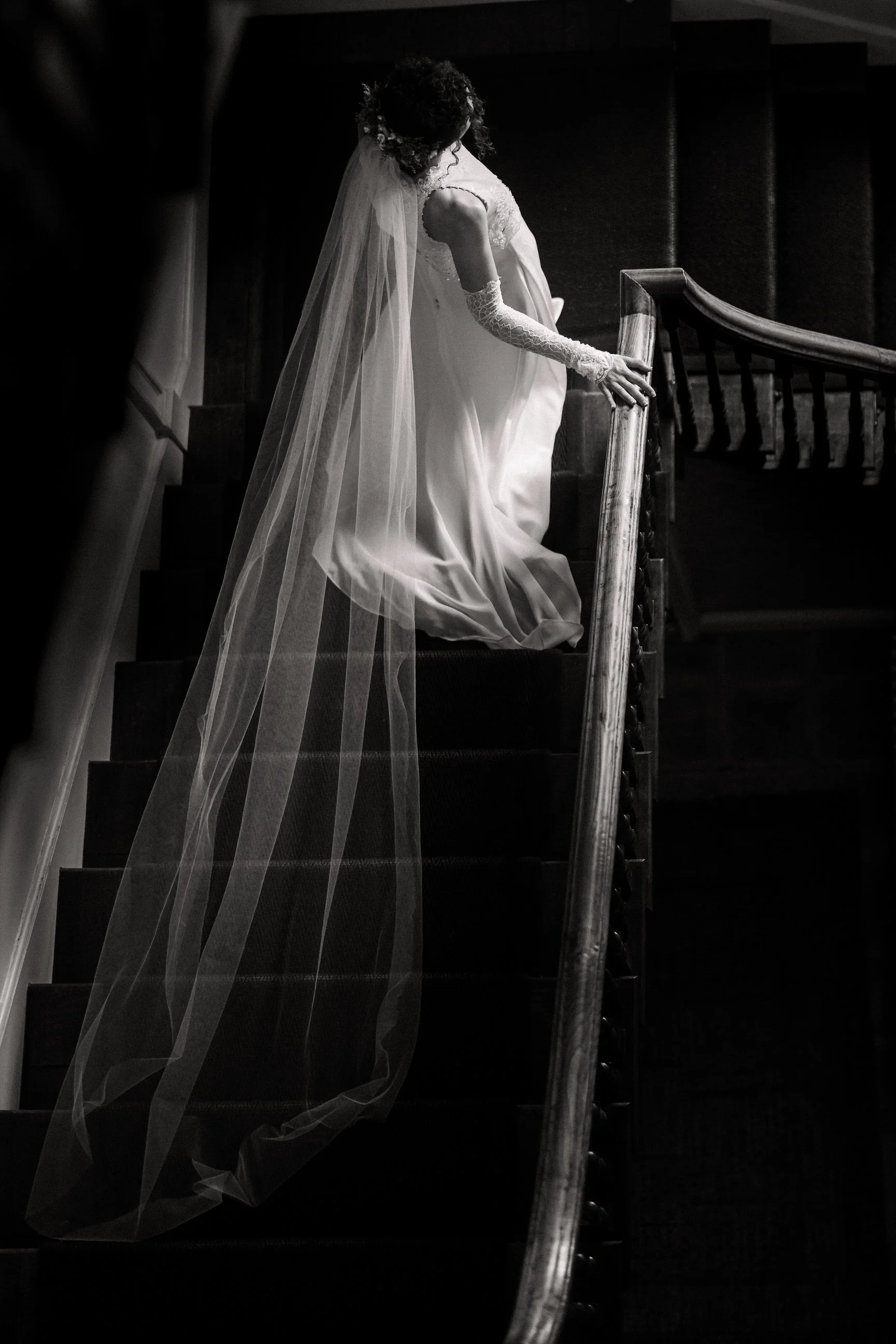 Black-and-white photograph of a bride in a wedding gown, long veil, and lace gloves ascending a staircase.