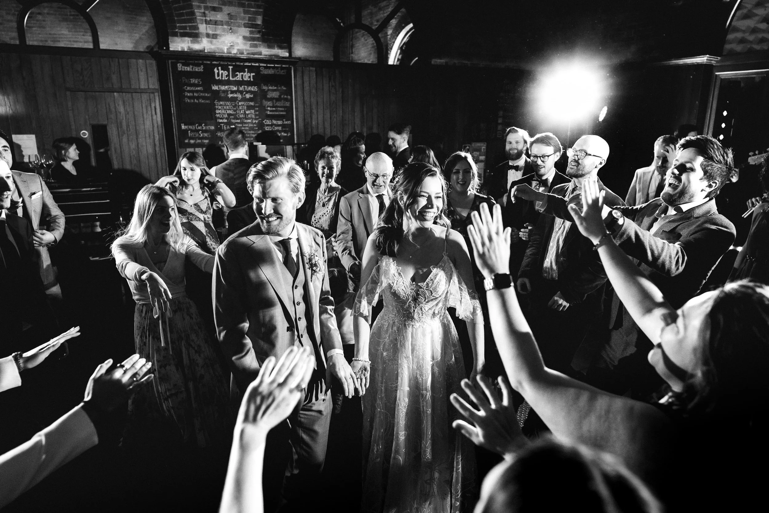 Black-and-white wedding reception photo of a bride and groom surrounded by cheering guests in a pub.