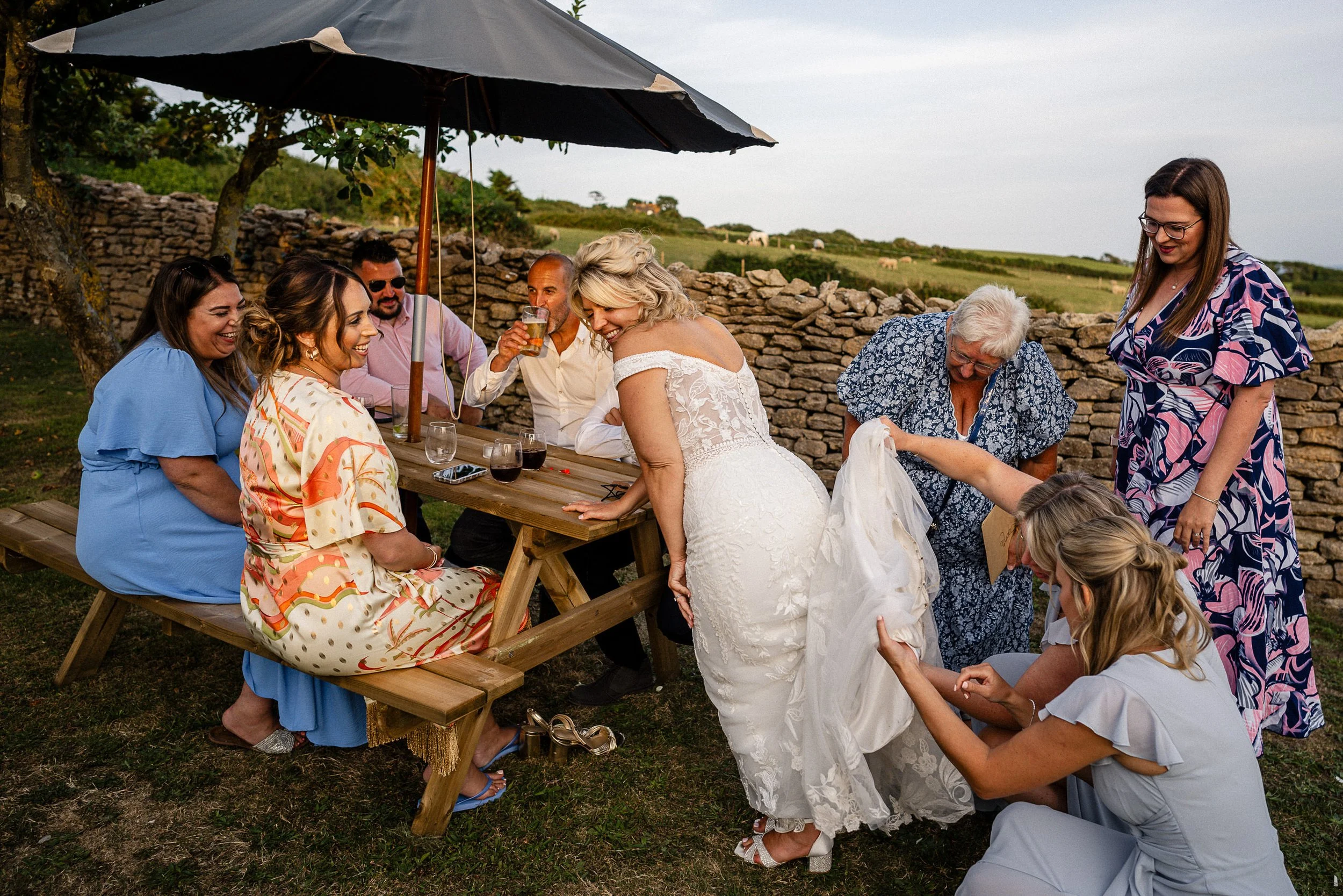 Bride in a lace wedding dress celebrating with friends and family at an outdoor picnic table in the countryside
