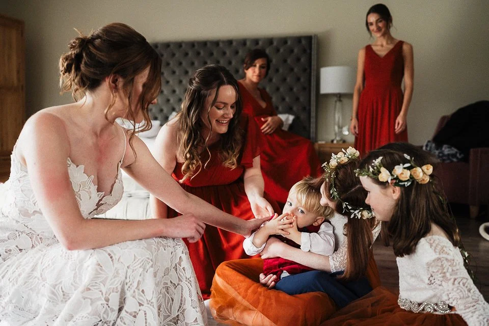 Bride in a lace wedding dress and bridesmaids in red dresses prepare with children in a bedroom.