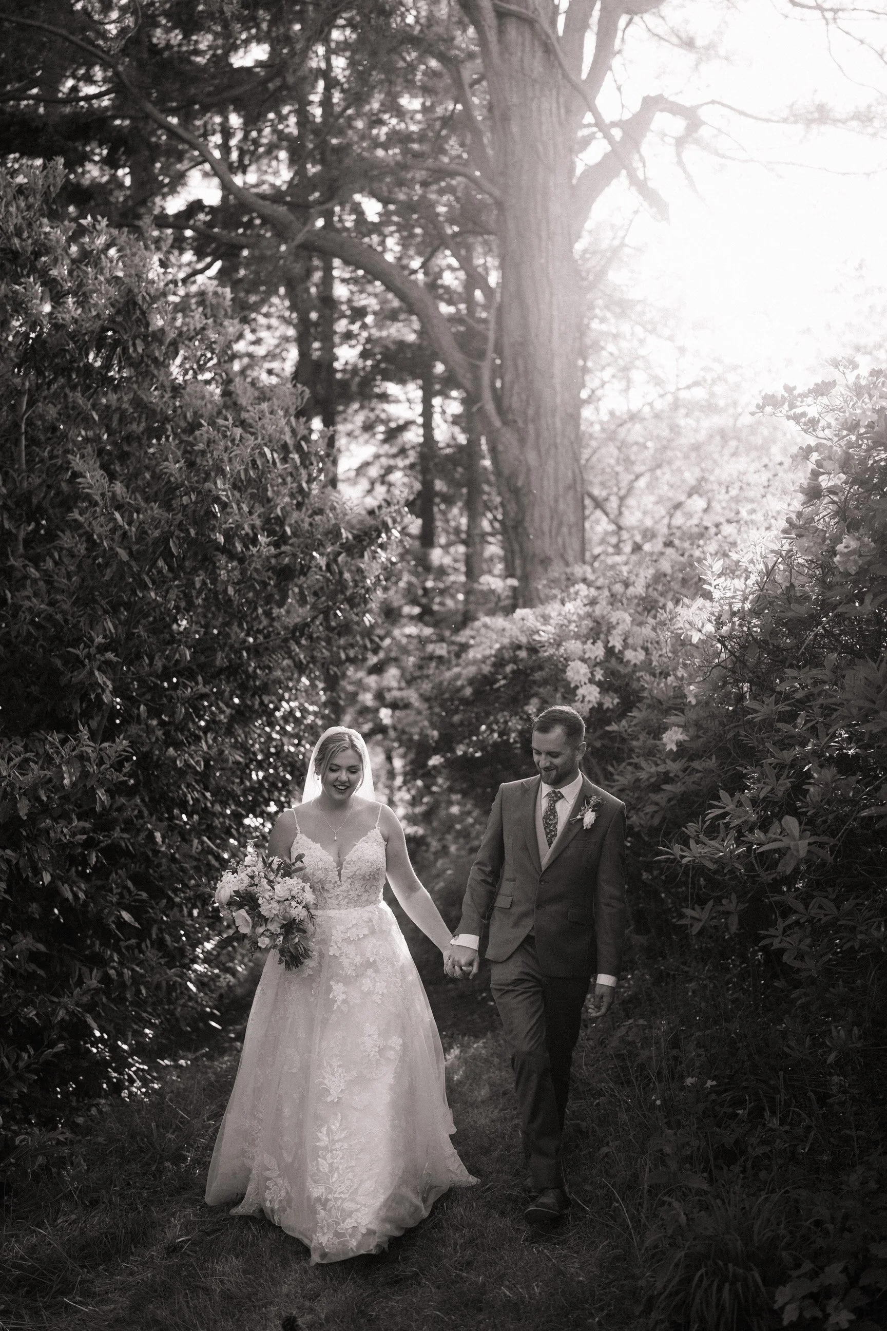 Black-and-white wedding portrait of a bride and groom holding hands while walking through a sunlit garden path.