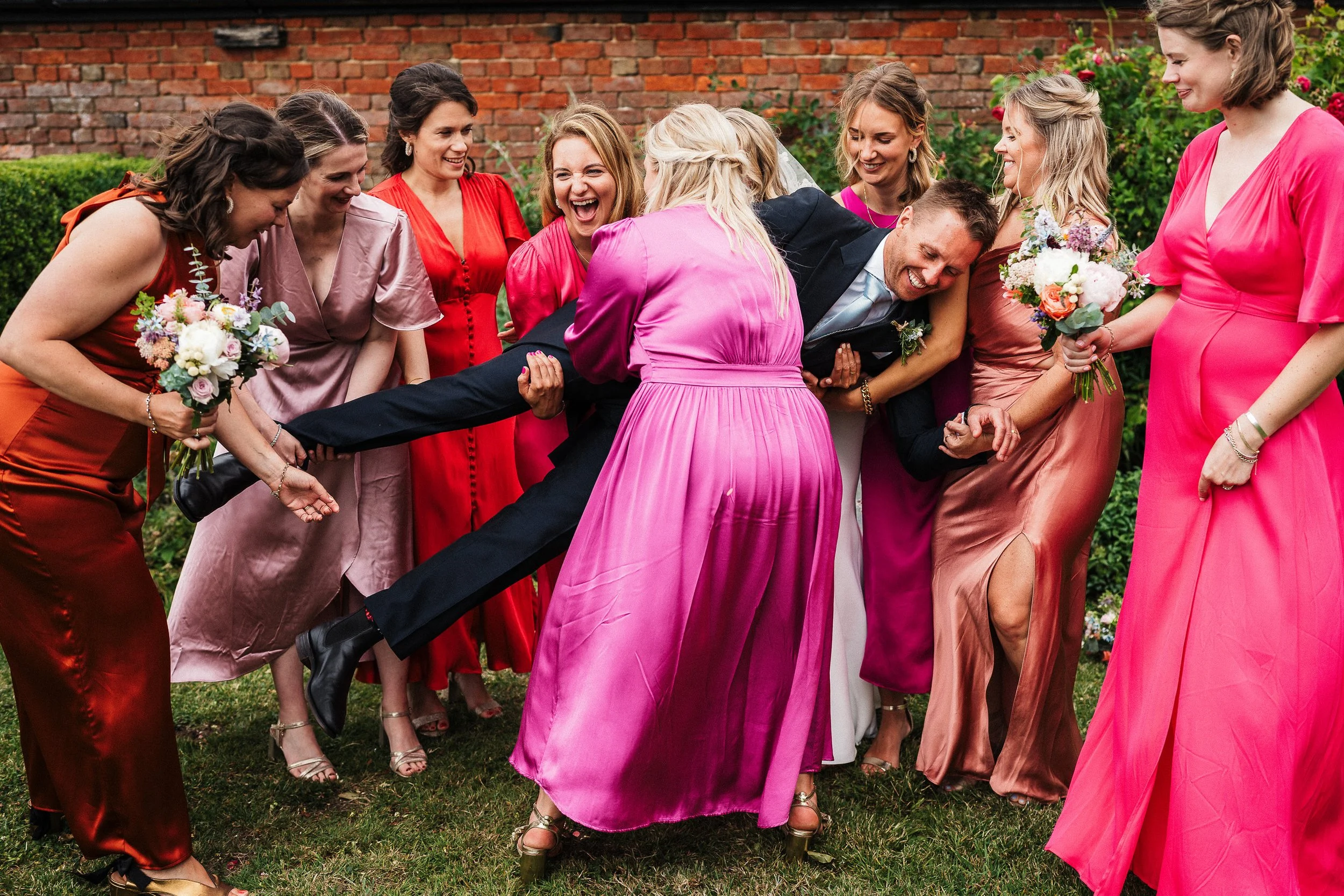 Wedding guests laughing and playfully lifting the groom outdoors while holding bouquets.