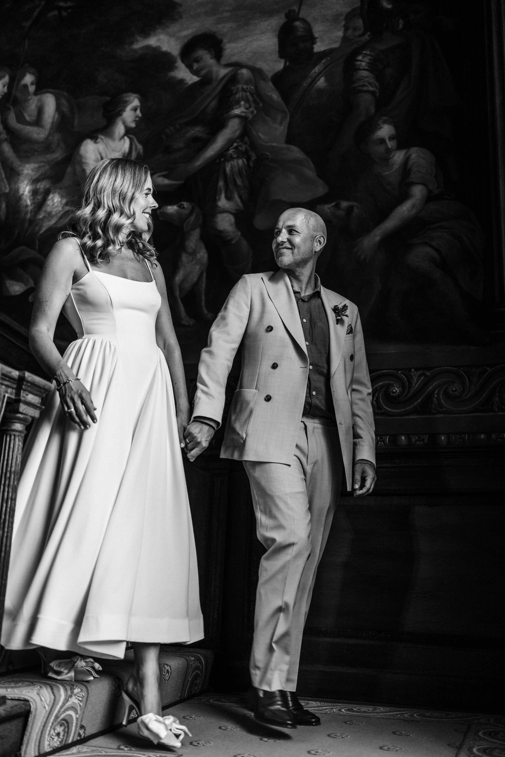 Black-and-white wedding portrait of a smiling bride and groom holding hands in front of a classical mural.