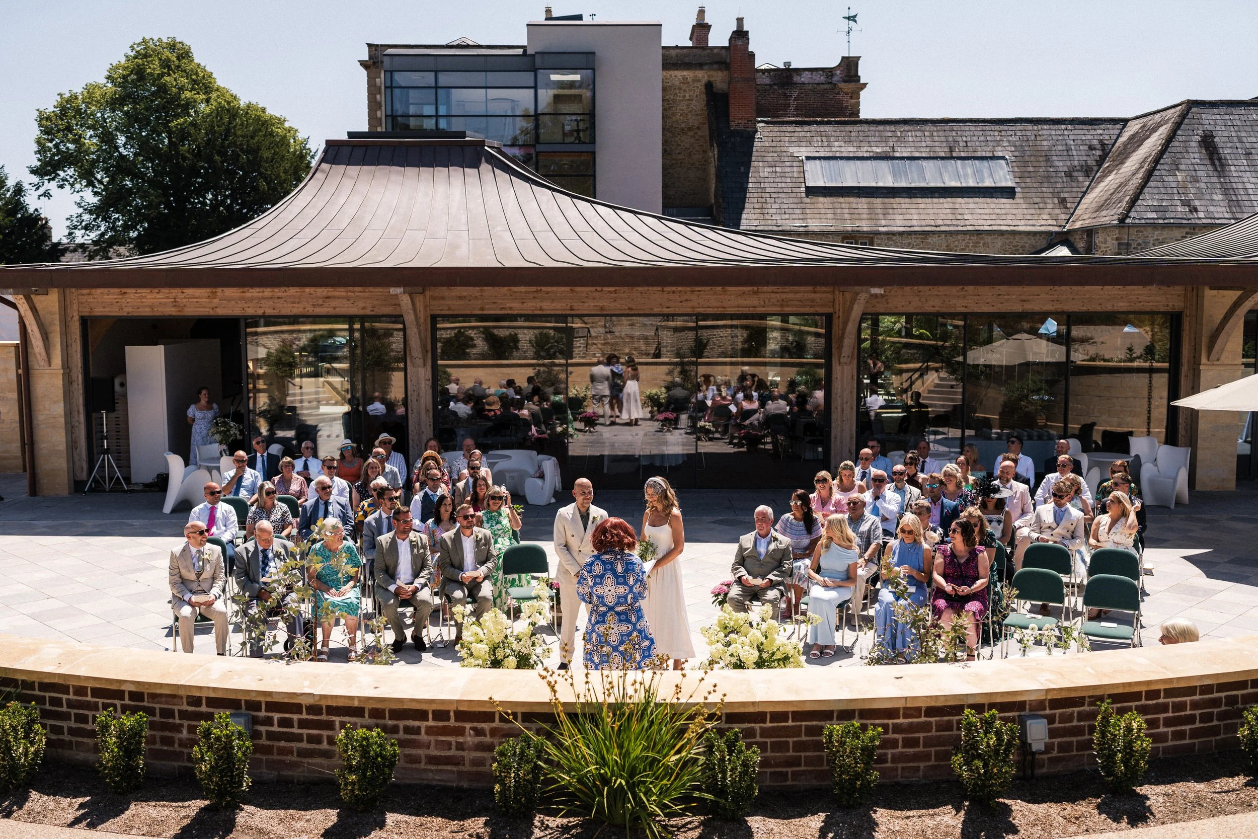 Outdoor wedding ceremony with bride, groom, officiant, and seated guests at a historic stone venue.
