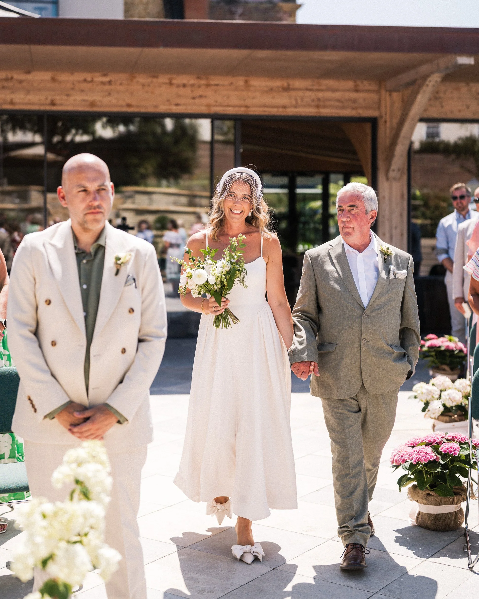 Bride carrying a white floral bouquet walks with her father at an outdoor wedding ceremony, accompanied by a groom in a cream suit.