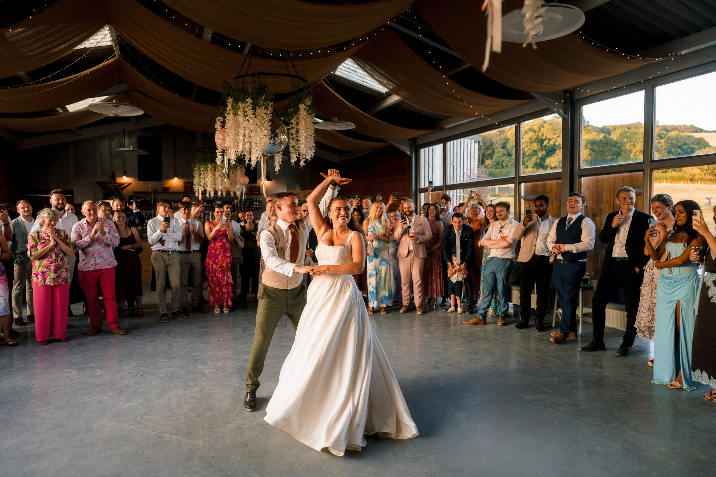 Bride and groom dance at a wedding reception as guests cheer and take photos in a decorated rustic venue.