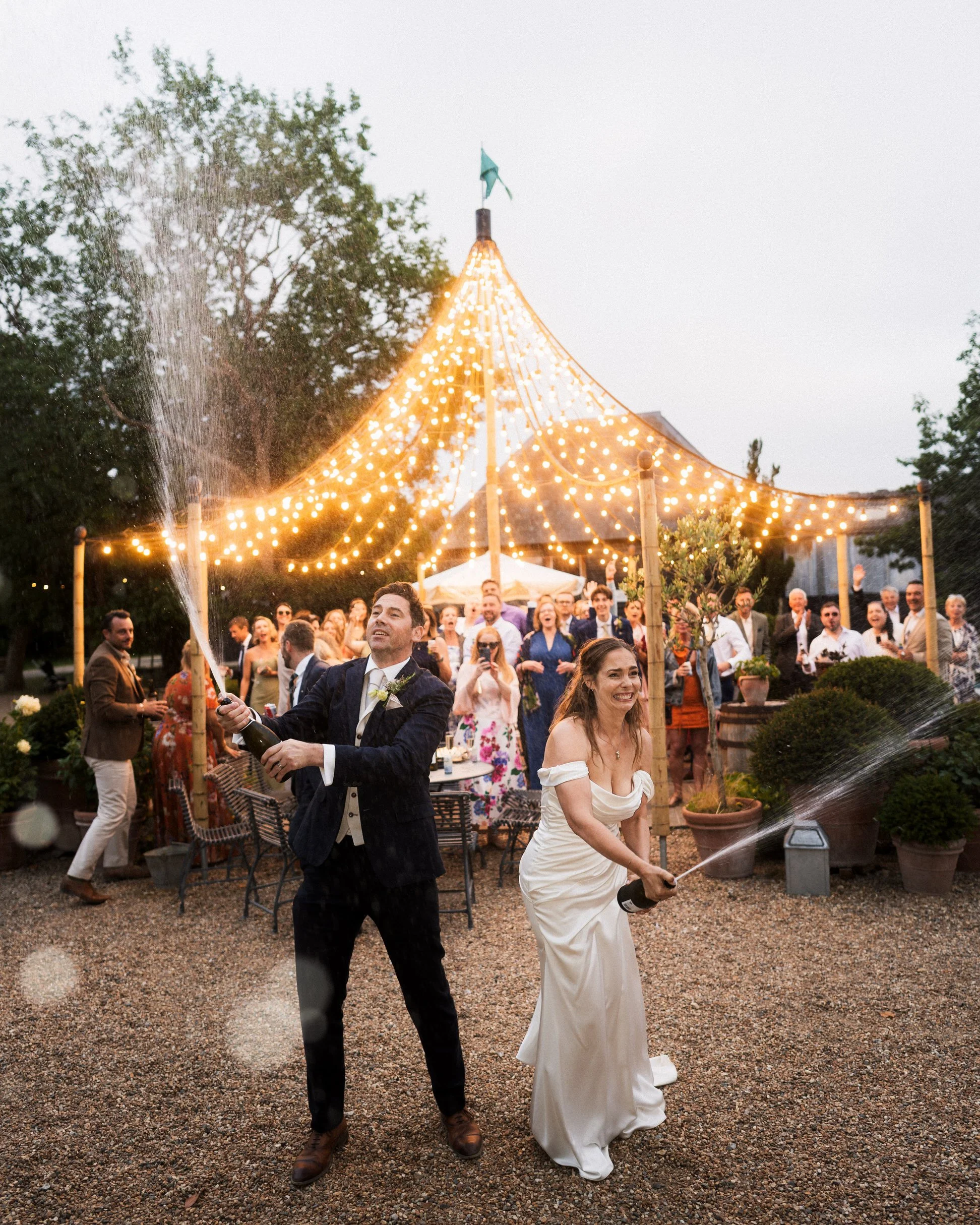 Newlywed couple sprays champagne at an outdoor wedding reception beneath glowing string lights as guests celebrate