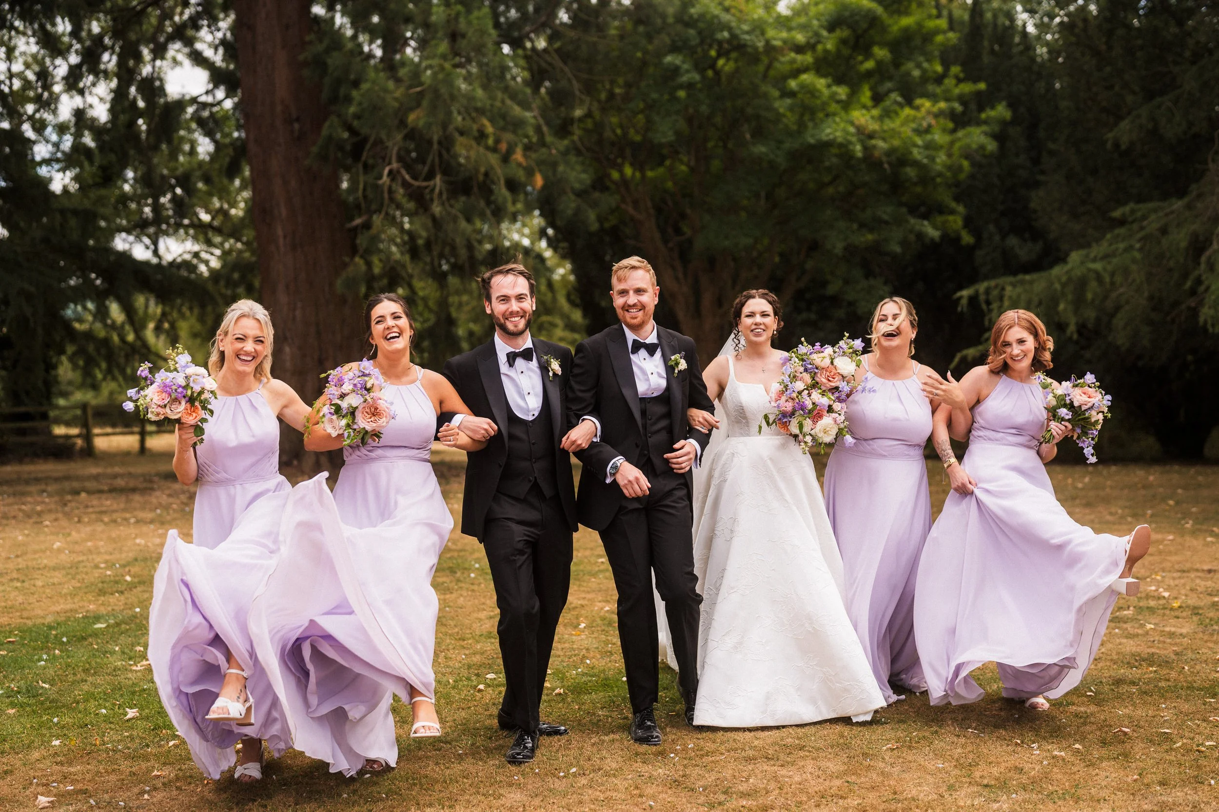 Joyful bride, grooms, and bridesmaids in lavender dresses posing with bouquets outdoors at a wedding.