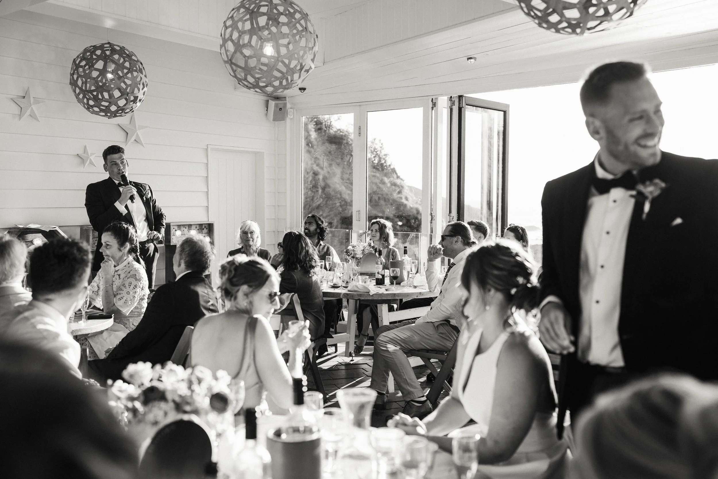 Black-and-white wedding reception with guests dining while a man in a tuxedo gives a speech