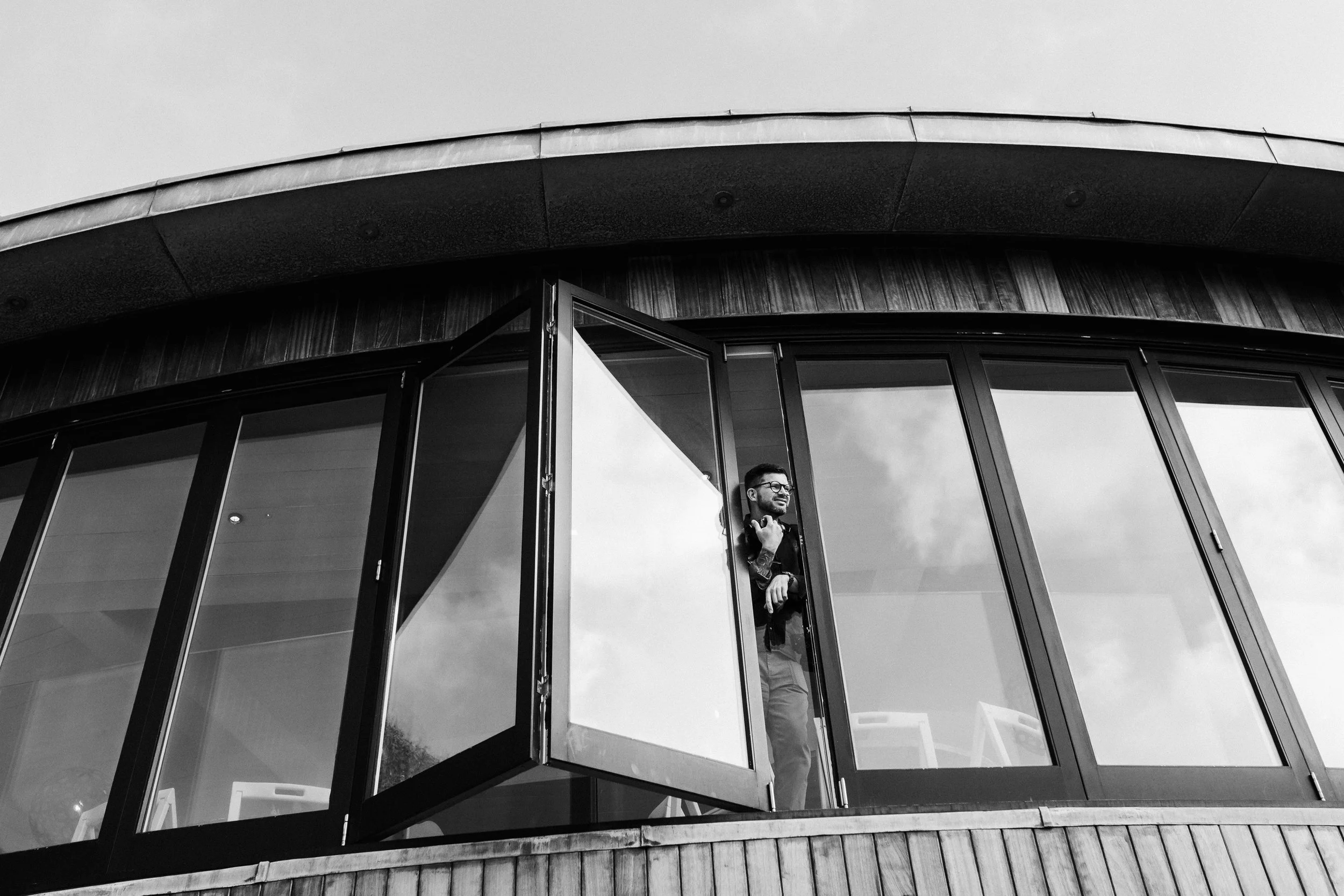 A man standing at an open window on a modern curved building with glass walls, smiling and looking outside.
