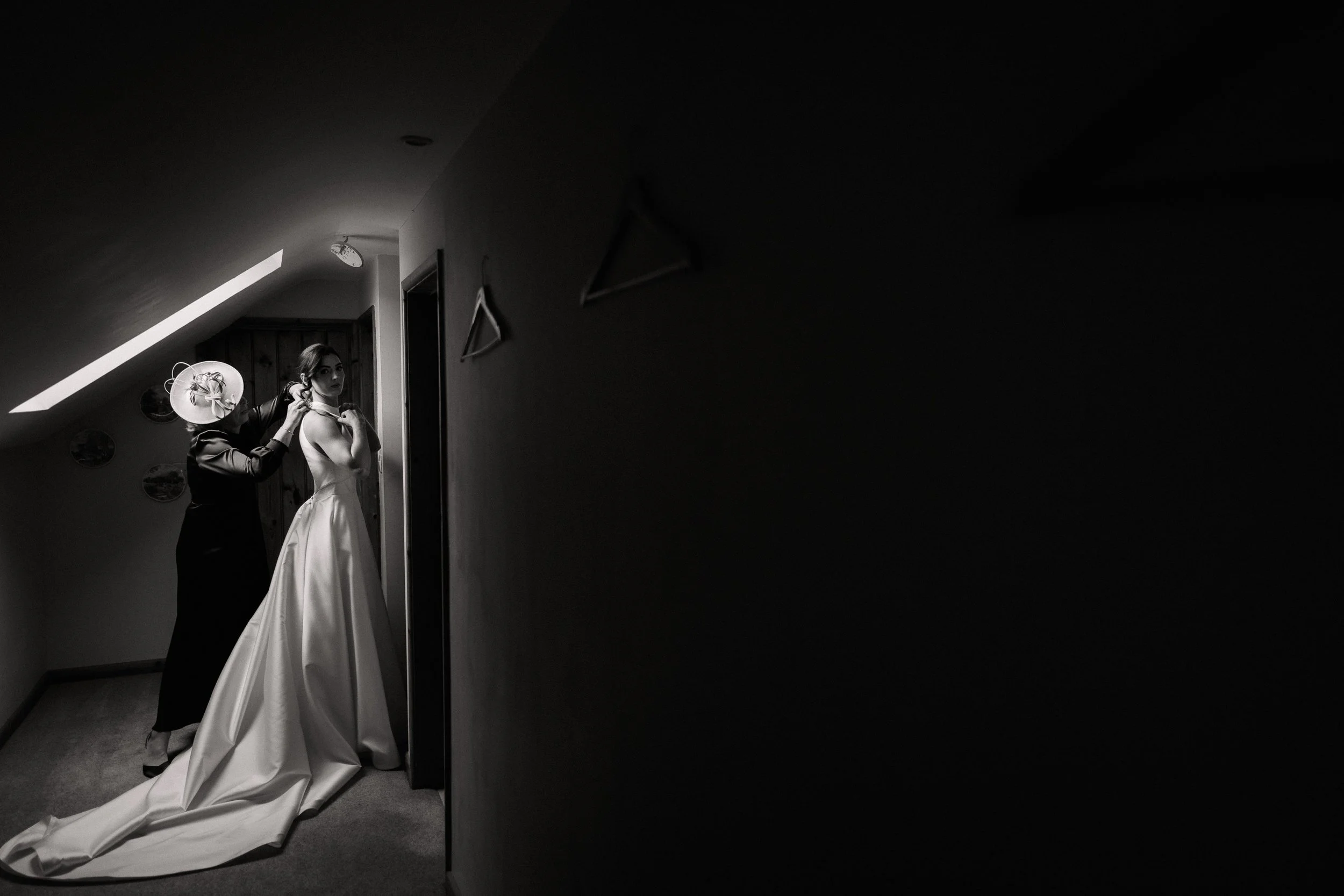 A bride in a wedding dress having her earring fitted by a woman with a hat in a dimly lit hallway.