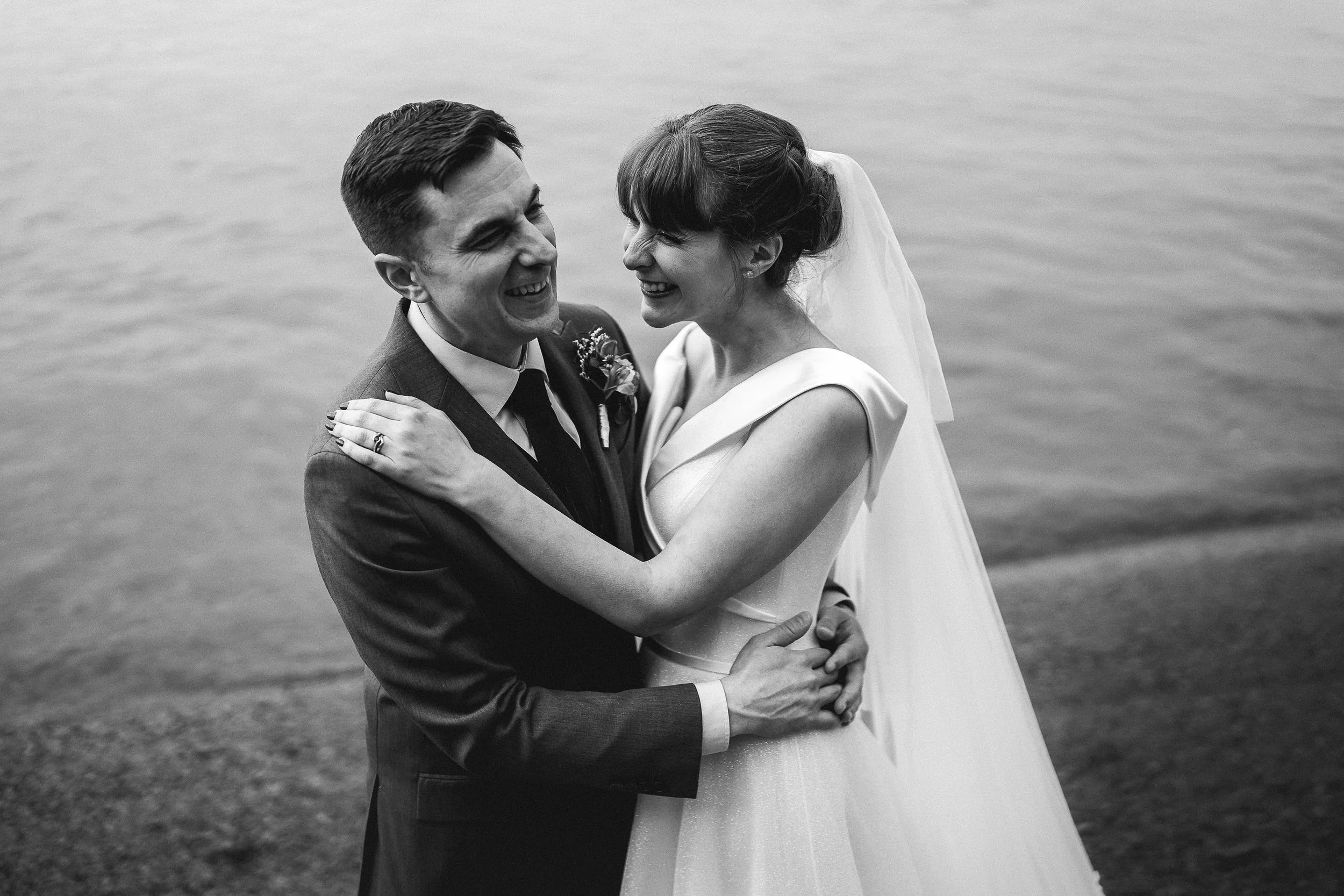 Black-and-white wedding portrait of a laughing bride and groom embracing beside the water