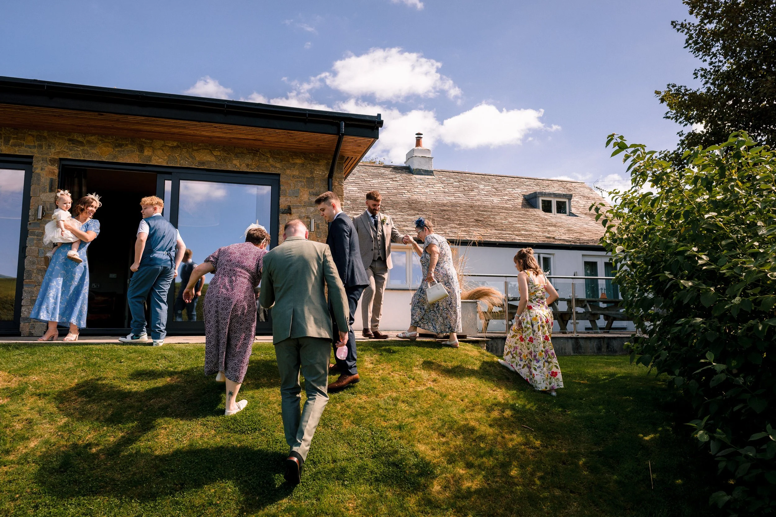 Wedding guests gather on a sunny lawn outside a stone house.