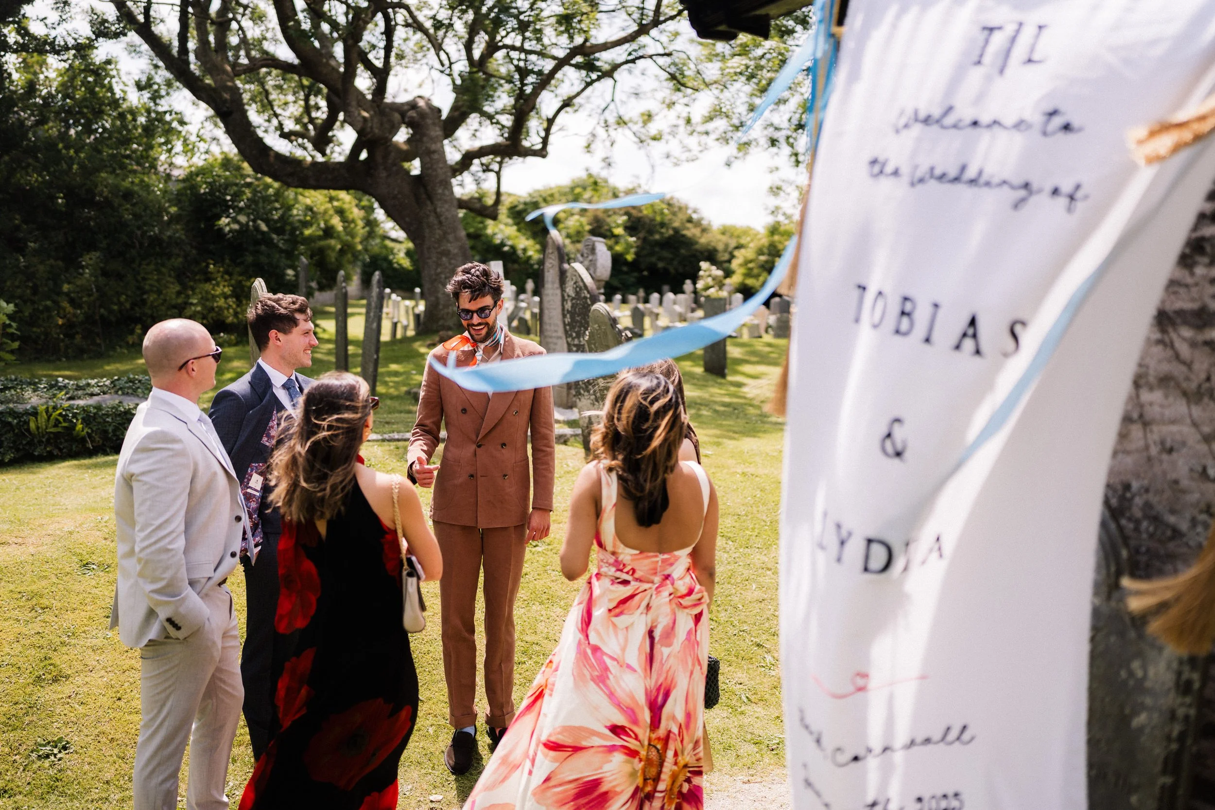 Wedding guests gather in a cemetery beside a handwritten welcome sign decorated with blue ribbons.