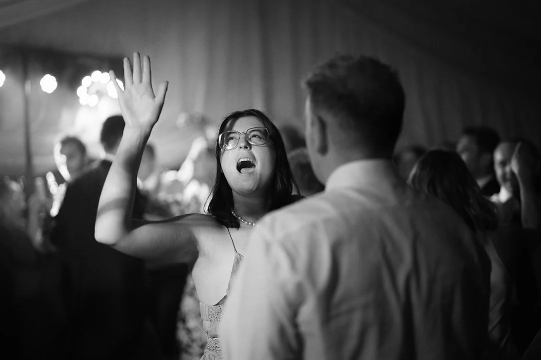 Black-and-white photo of a woman wearing glasses raising her hand and singing at a crowded wedding reception.