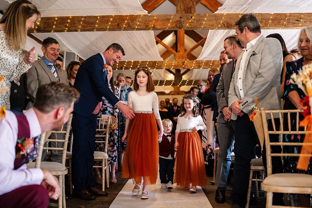 Children walk down the aisle at a rustic barn wedding surrounded by guests and string lights