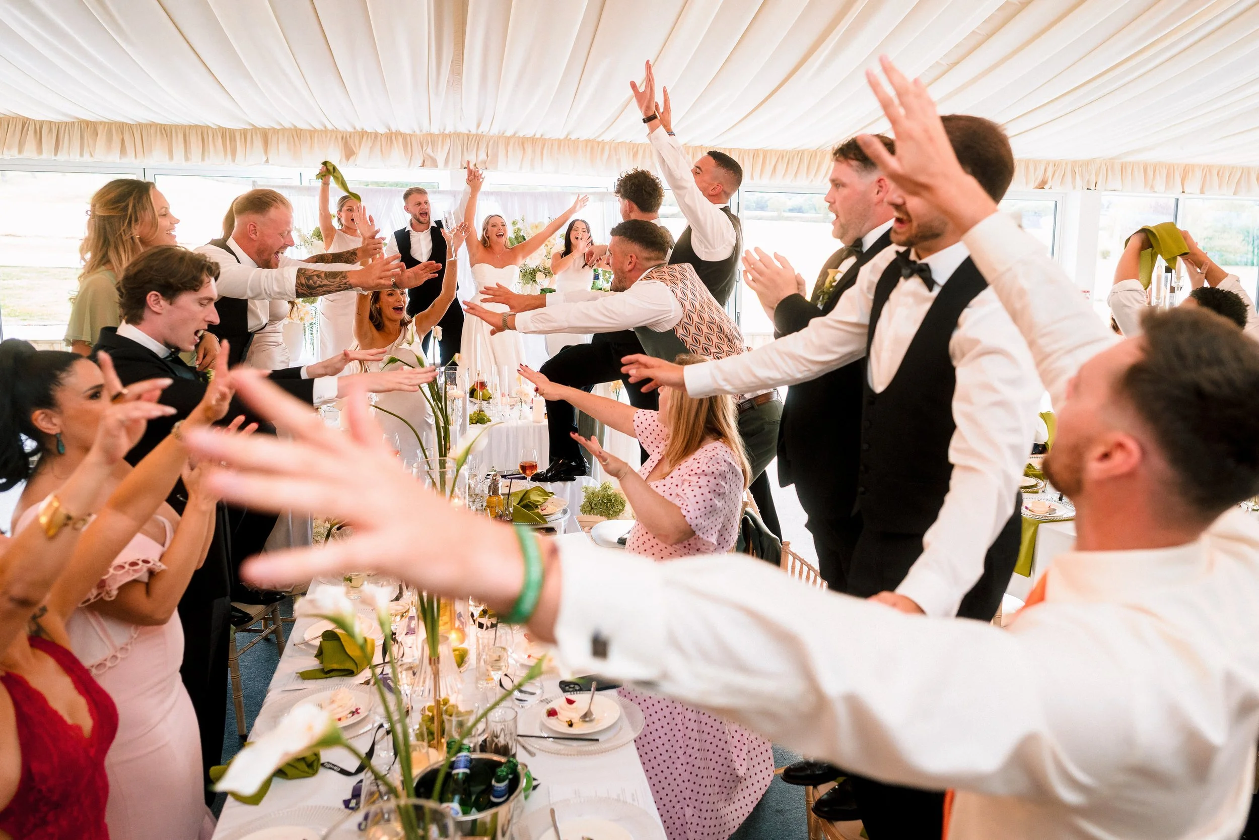 Wedding guests celebrating and dancing around a decorated reception table beneath a white marquee tent