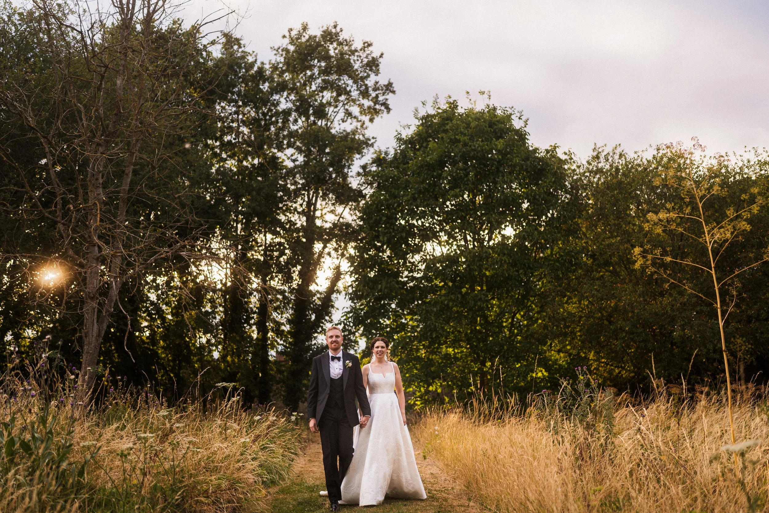 Newlywed couple walking hand in hand through a sunlit meadow surrounded by trees