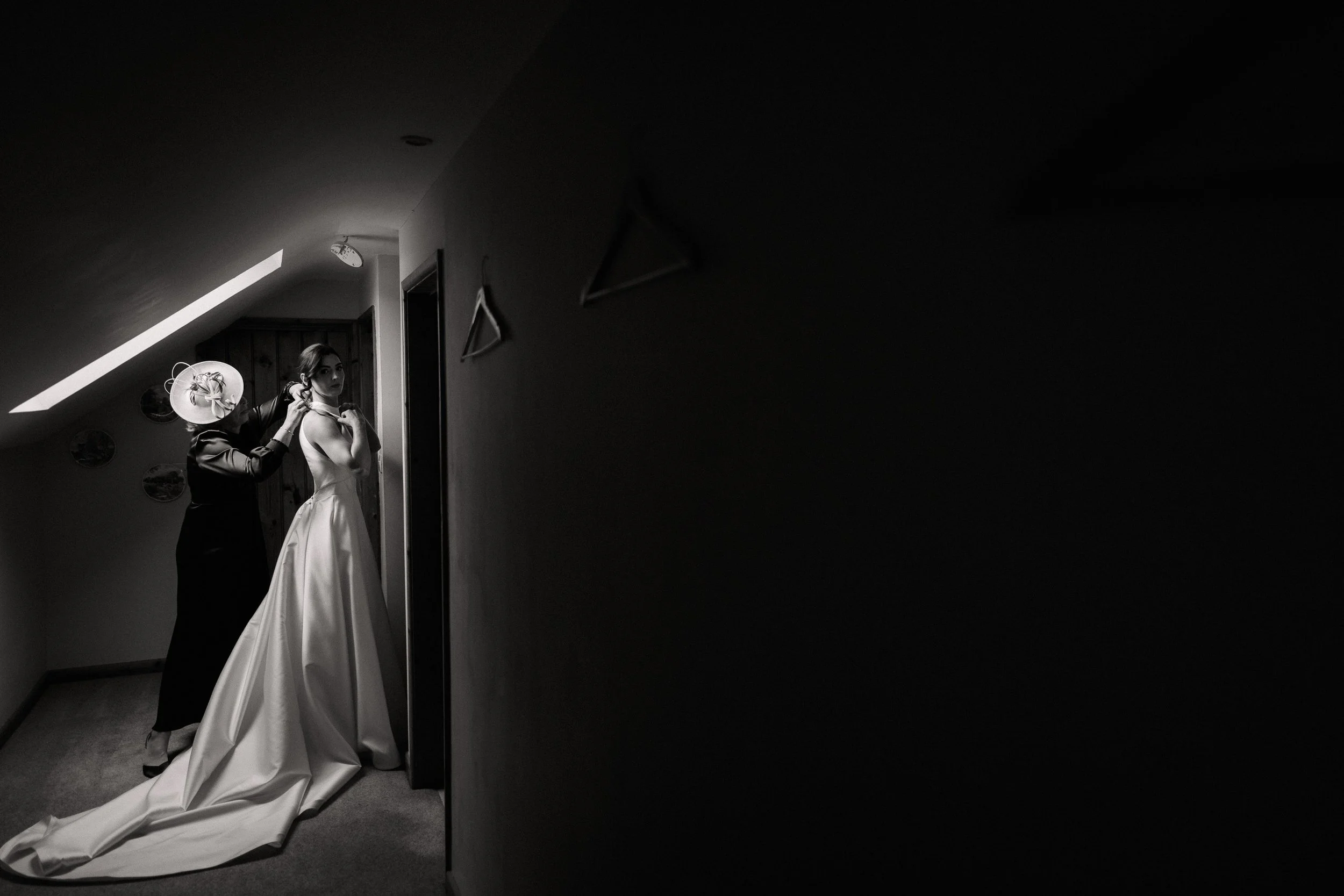 Black-and-white photo of a bride in a long satin wedding gown being helped by another woman indoors.