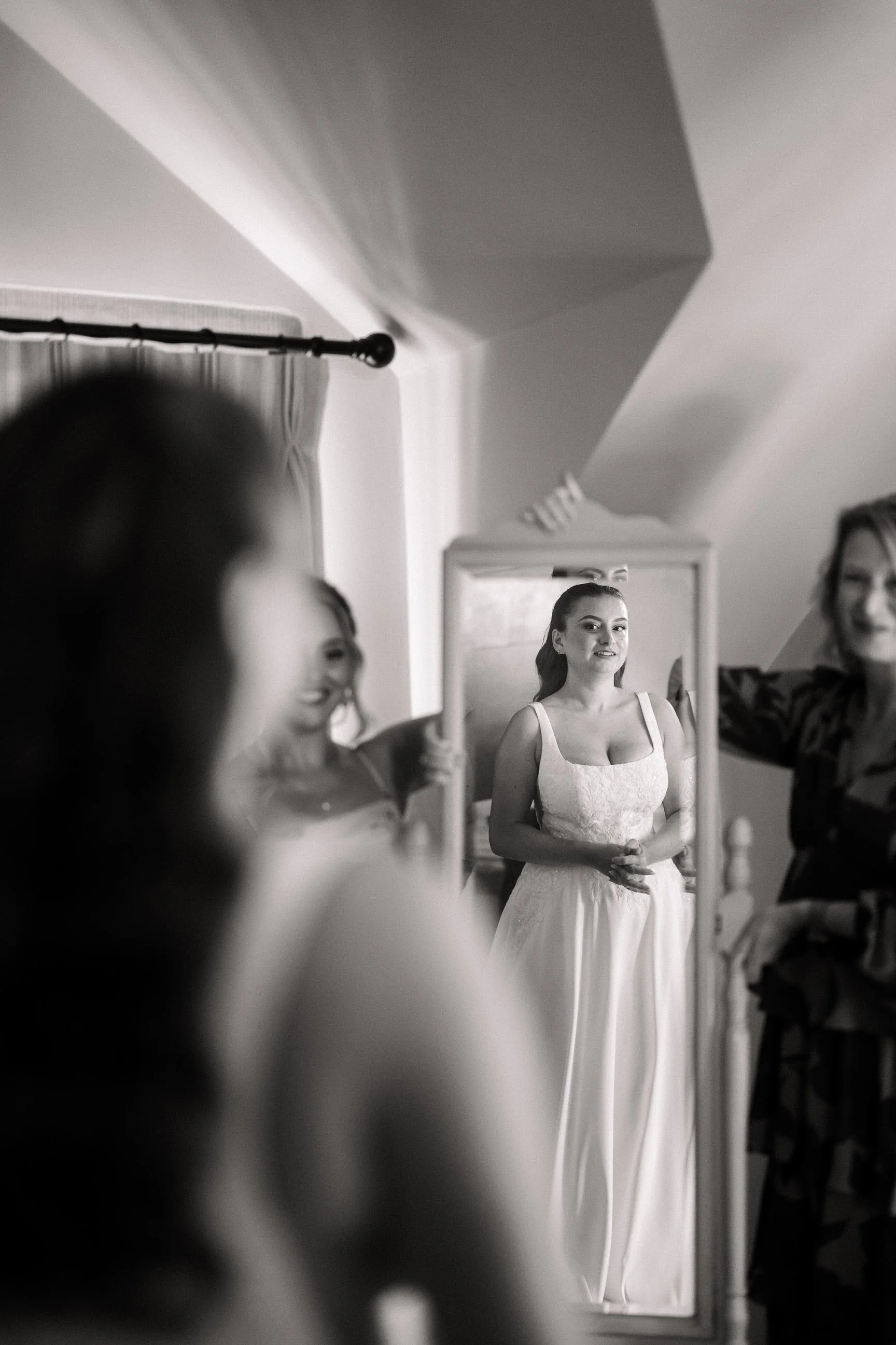 Black-and-white photo of a bride in a white dress standing before a mirror while people help her prepare.