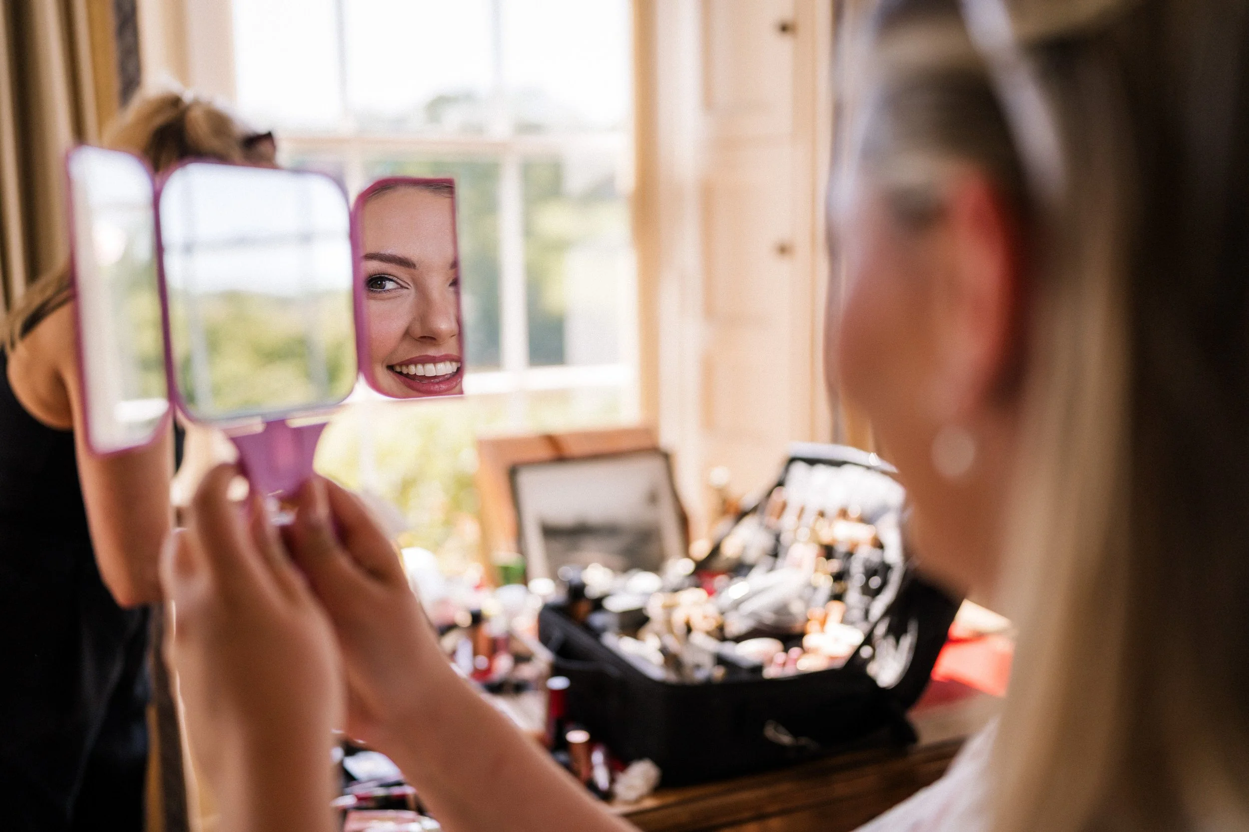 Smiling woman applying makeup while looking into a handheld mirror