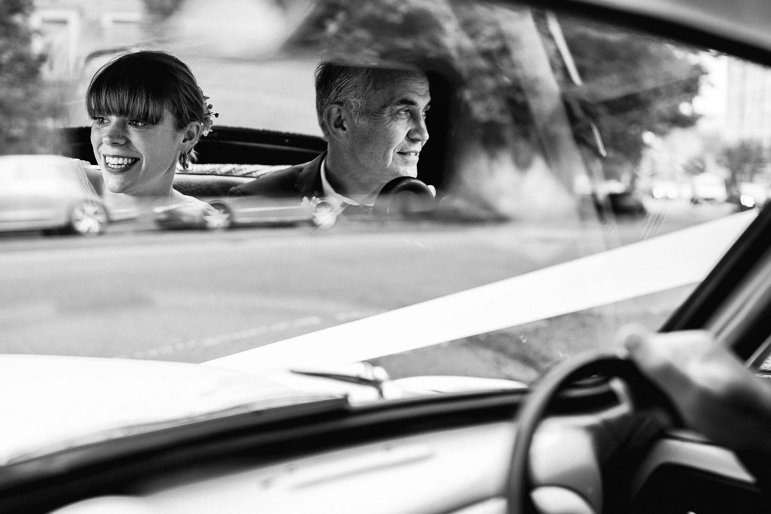 Black-and-white photo of a smiling bride and groom riding in a vintage car.