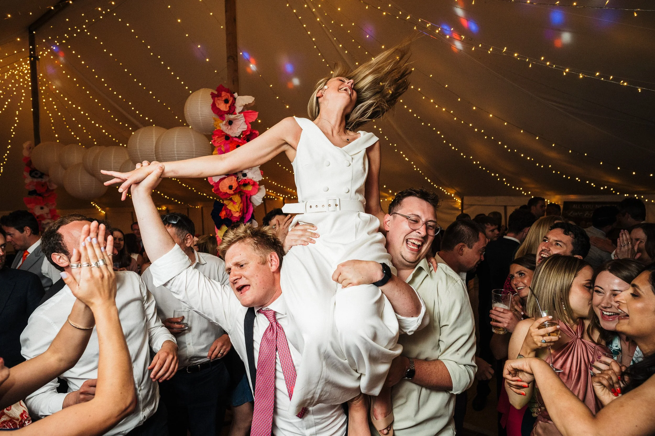 Joyful wedding guests lift a laughing bride in a white dress beneath string lights and colorful floral decorations.