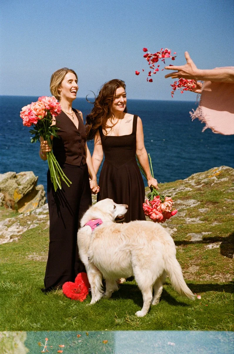 Two women holding hands during a wedding ceremony outdoors, with a scenic ocean view in the background. One woman is holding a bouquet of pink flowers, and a golden retriever is standing in front of them.