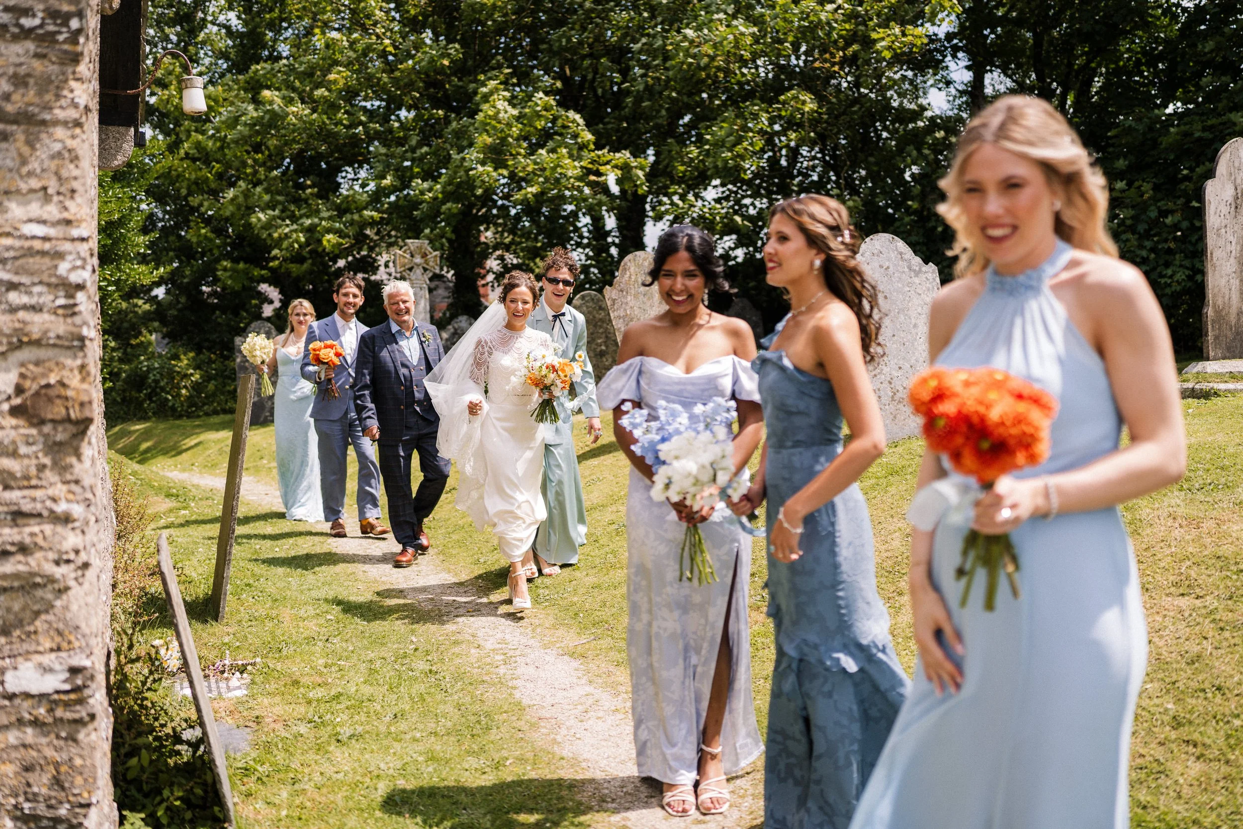 Bride, groom, and wedding party walking through a cemetery with flower bouquets