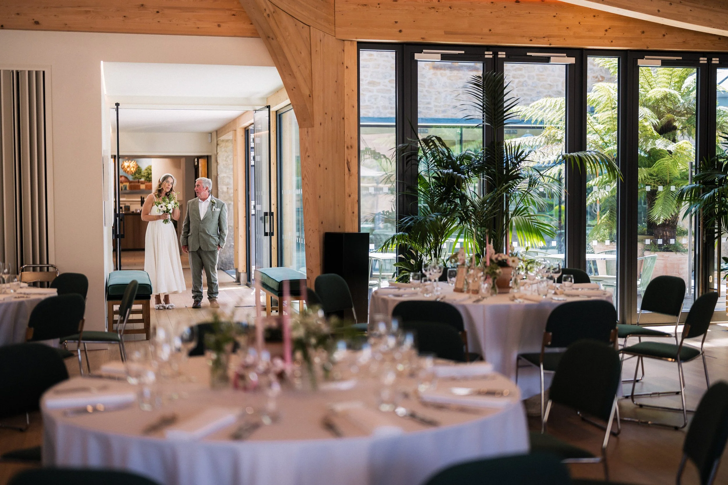 Bride and groom entering a sunlit wedding reception venue with decorated dining tables and tropical plants.