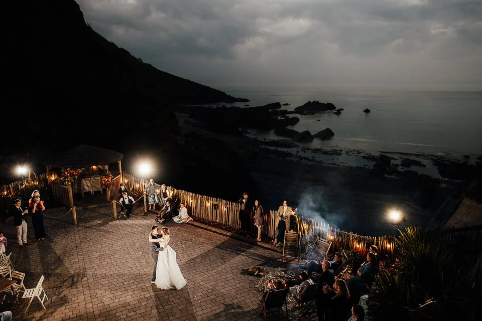 Bride and groom dancing at an outdoor seaside wedding reception overlooking rocky coastal cliffs at dusk.
