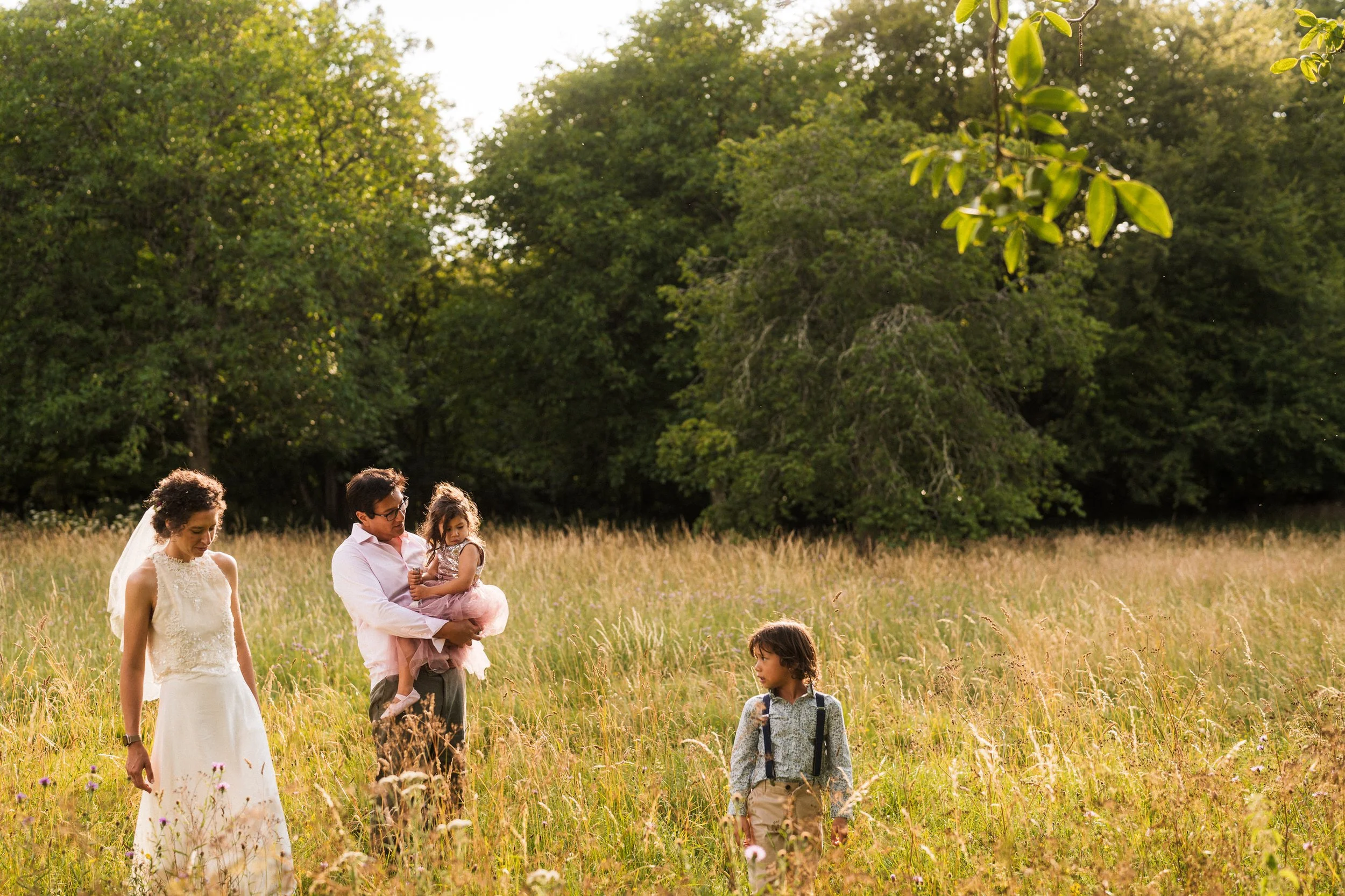 Bride, groom holding their daughter, and a young boy walking through a sunlit meadow bordered by trees