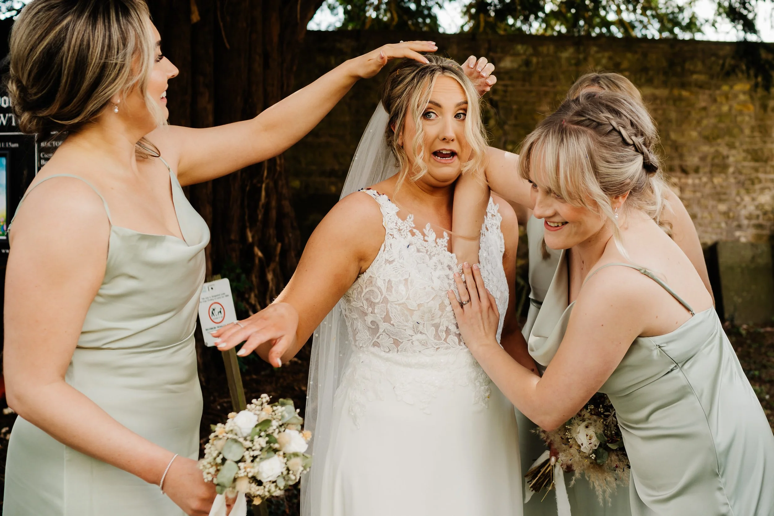 Bride reacting as two bridesmaids adjust her veil and hair outdoors