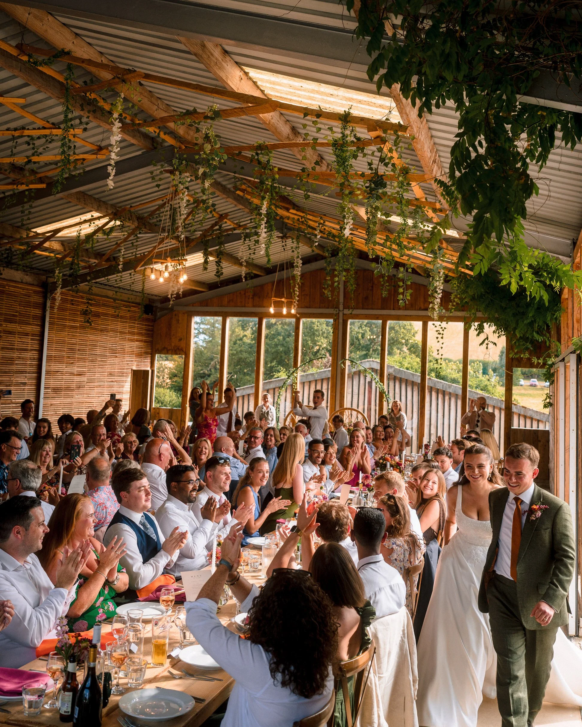 Wedding reception in a rustic barn, with guests applauding beneath greenery and wooden beams as the bride and groom stand together.