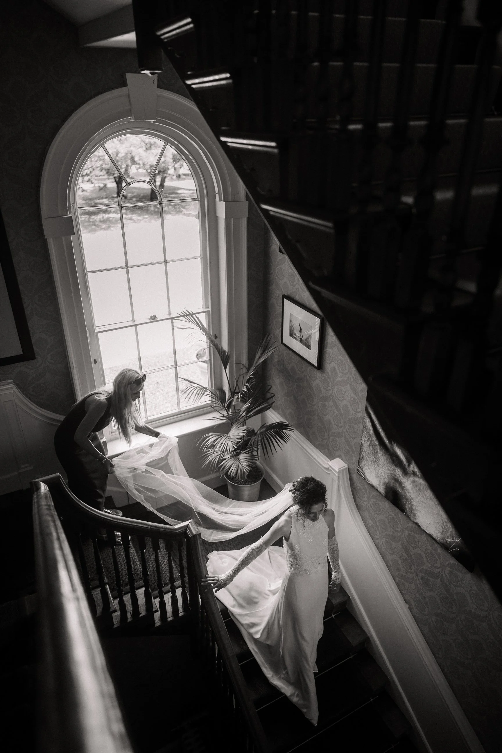 Black-and-white wedding photograph of a bride descending a grand staircase while a bridesmaid carries her cathedral-length veil.