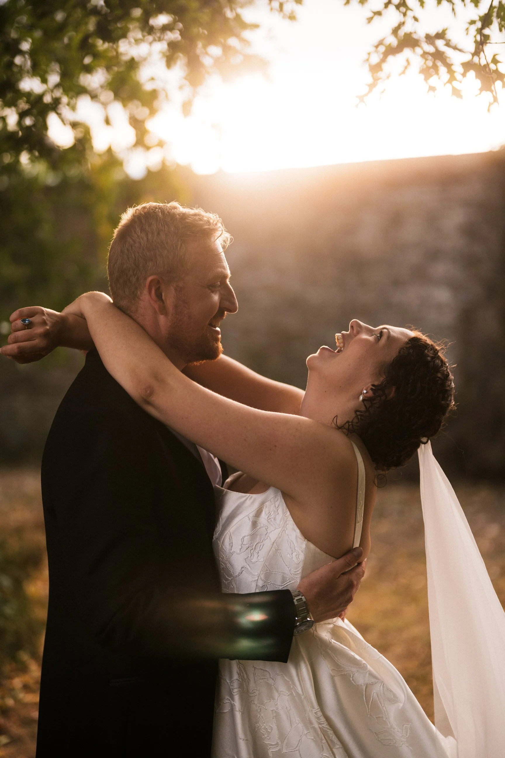 Newlywed couple laughing and embracing outdoors in warm sunset light, with the bride wearing a white wedding dress and veil.