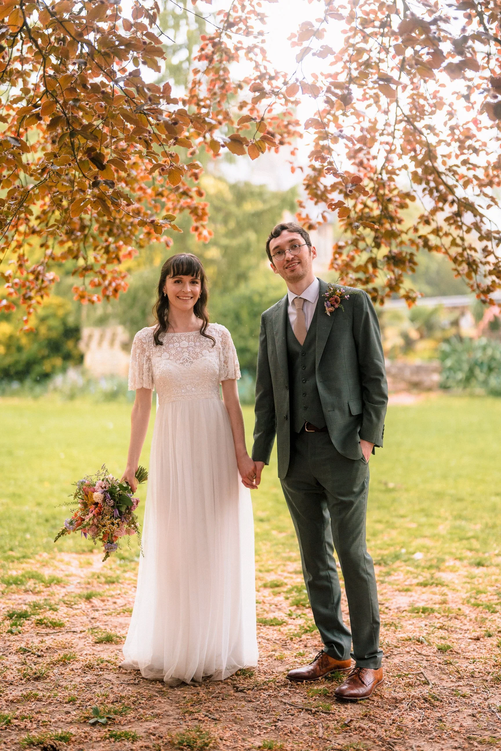 Newlywed couple holding hands outdoors beneath autumn trees, with the bride carrying a bouquet and the groom wearing a boutonniere.