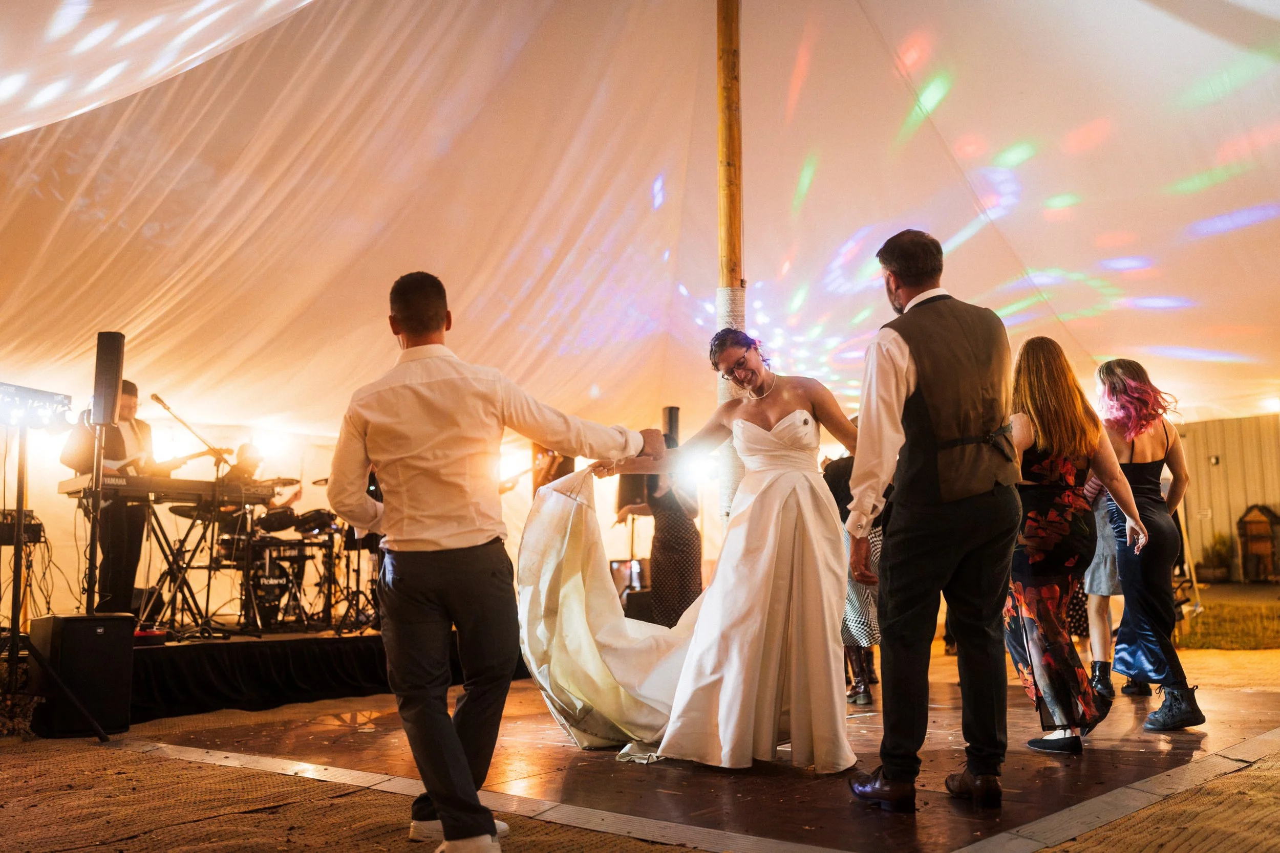 Bride and wedding guests dancing under colorful lights at a reception beside a live band