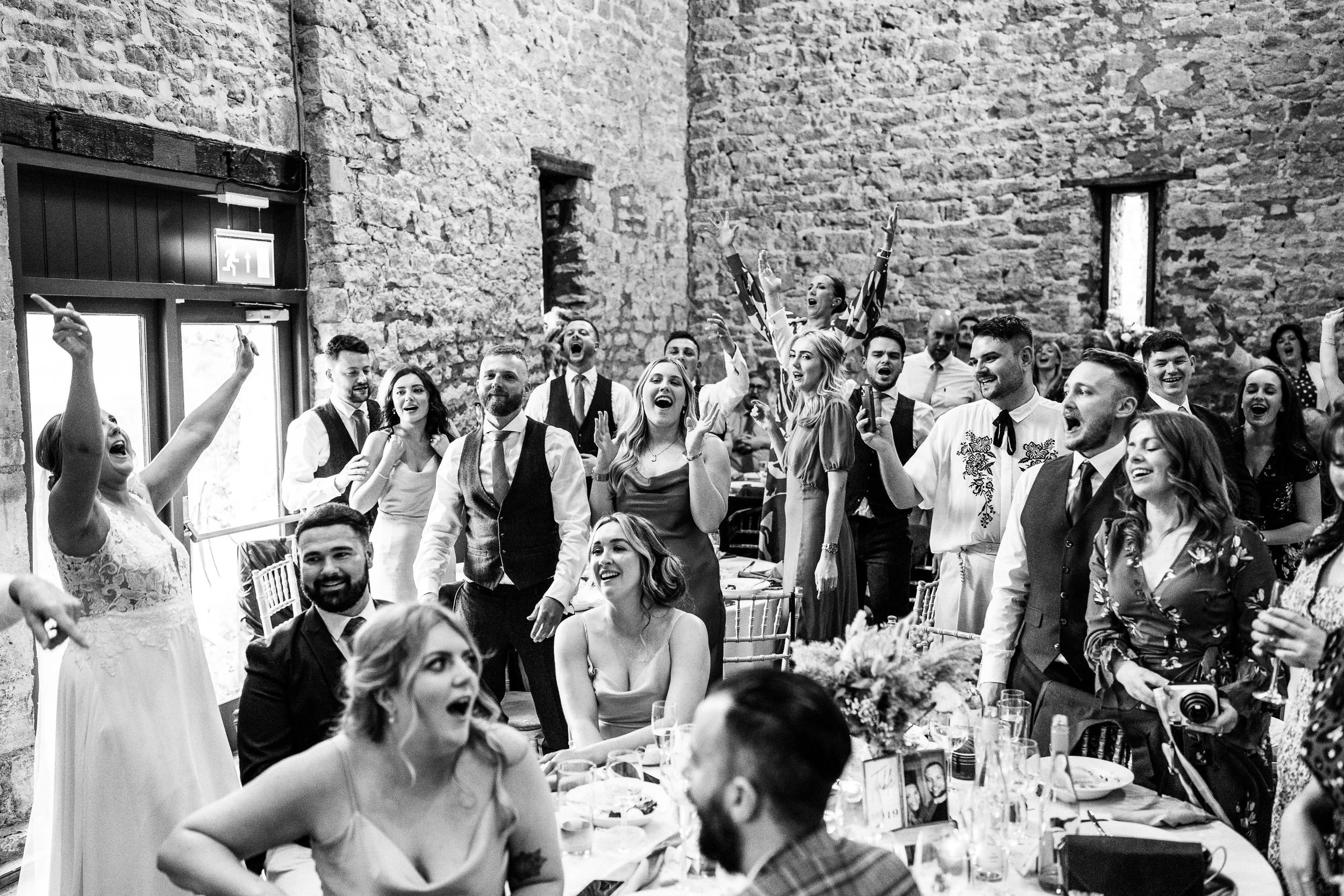 Black-and-white photo of wedding guests cheering and celebrating around a reception table in a rustic stone venue