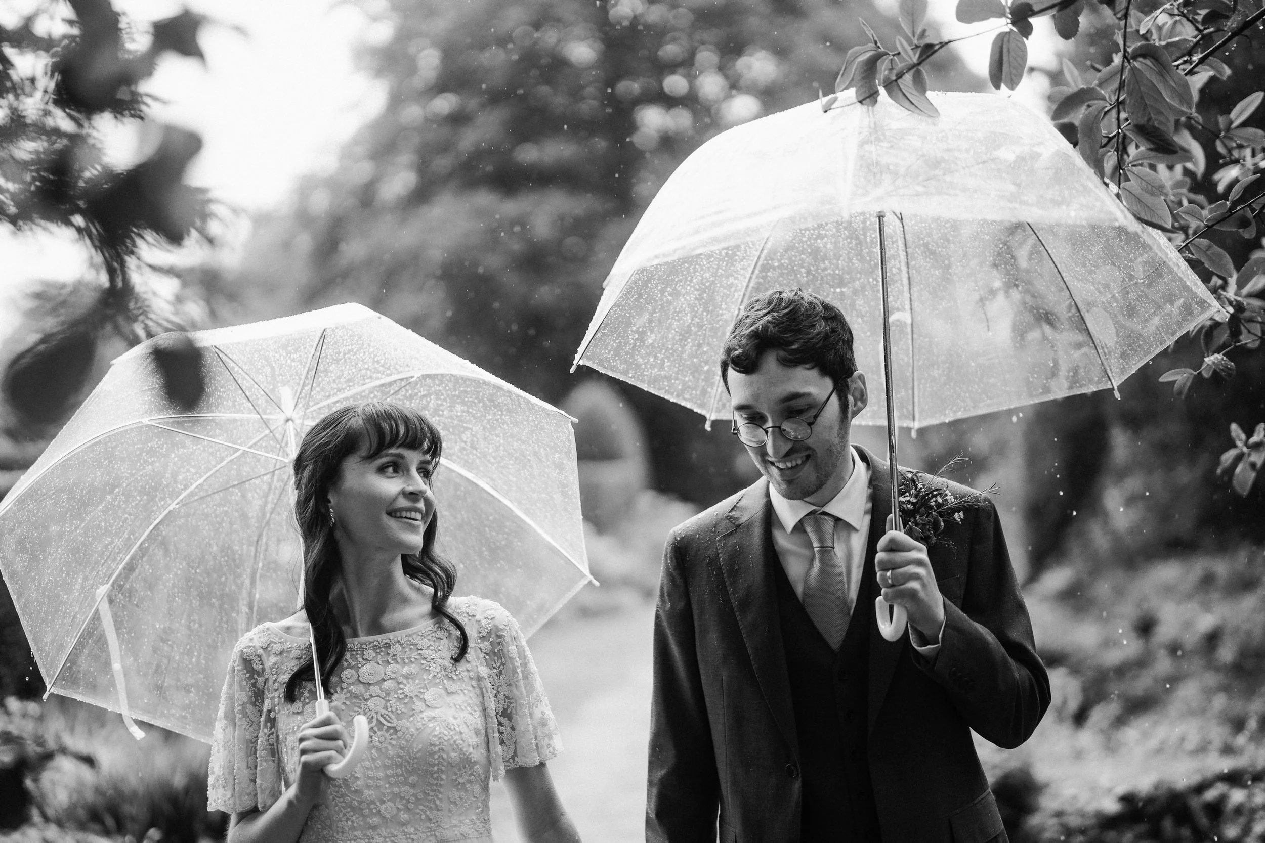 Black-and-white wedding portrait of a smiling bride and groom walking in the rain under clear umbrellas.