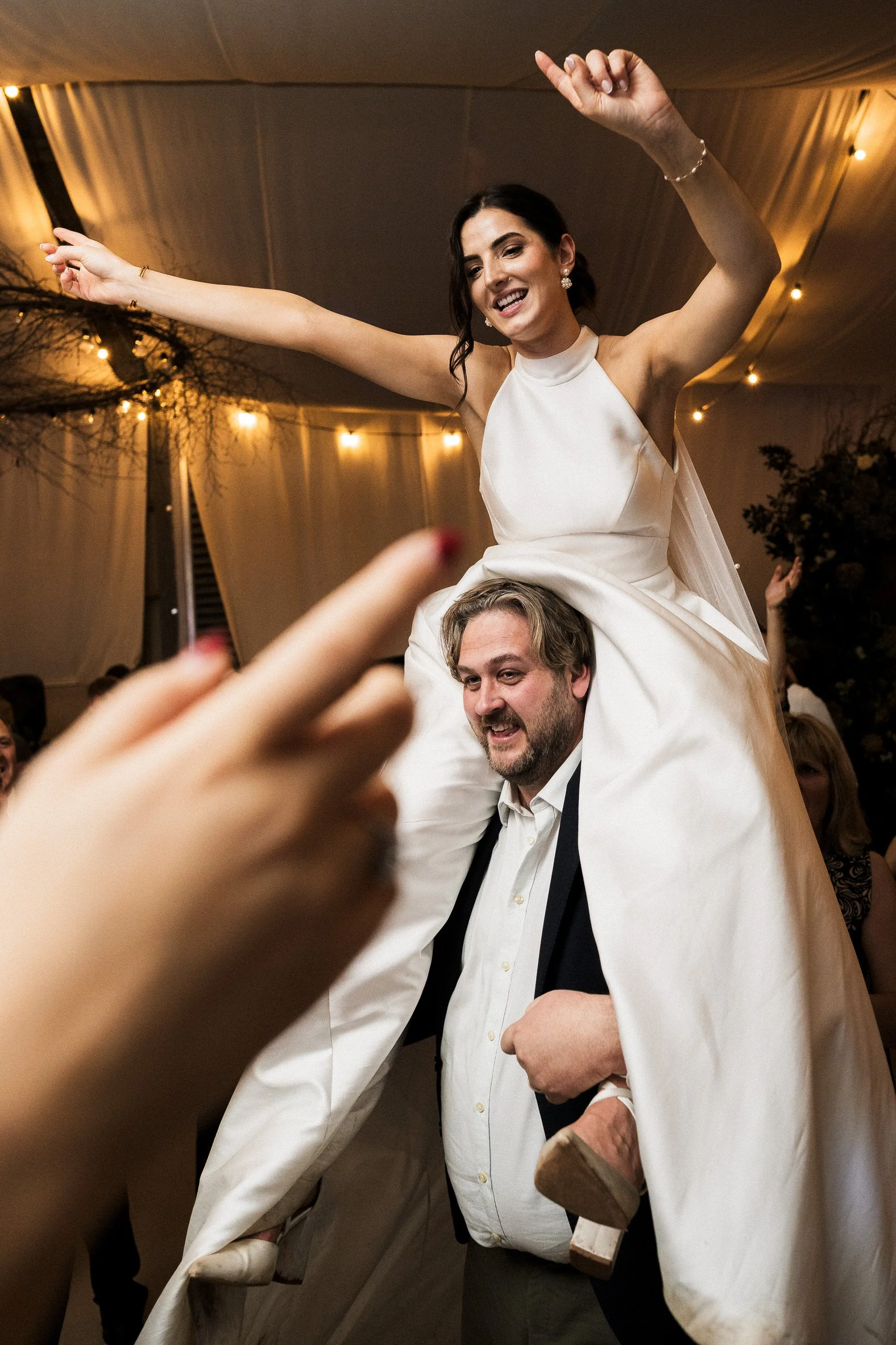 Bride with raised arms rides on a groom’s shoulders during a lively wedding reception under warm string lights.