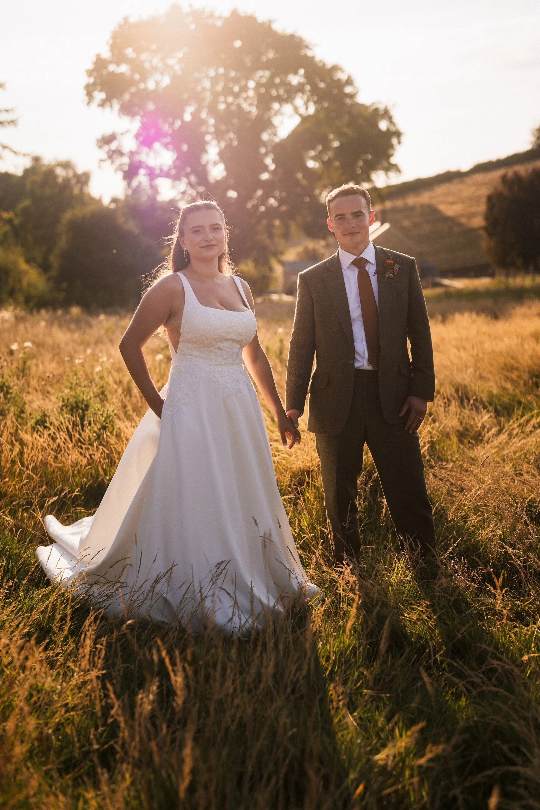 Newlywed couple holding hands in a sunlit meadow, wearing a white wedding dress and brown suit