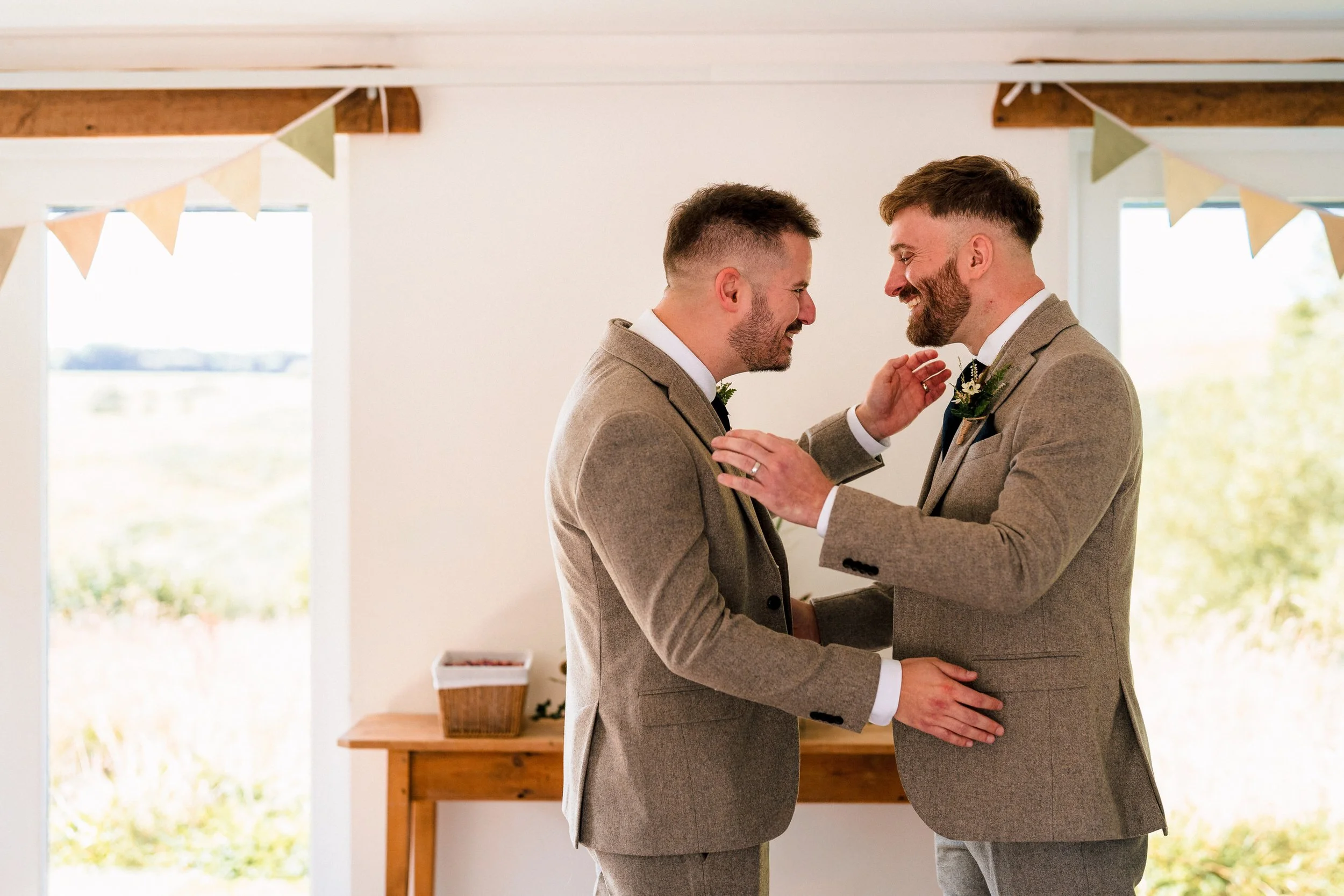 Two grooms in gray suits adjust each other’s boutonnieres at a wedding ceremony.