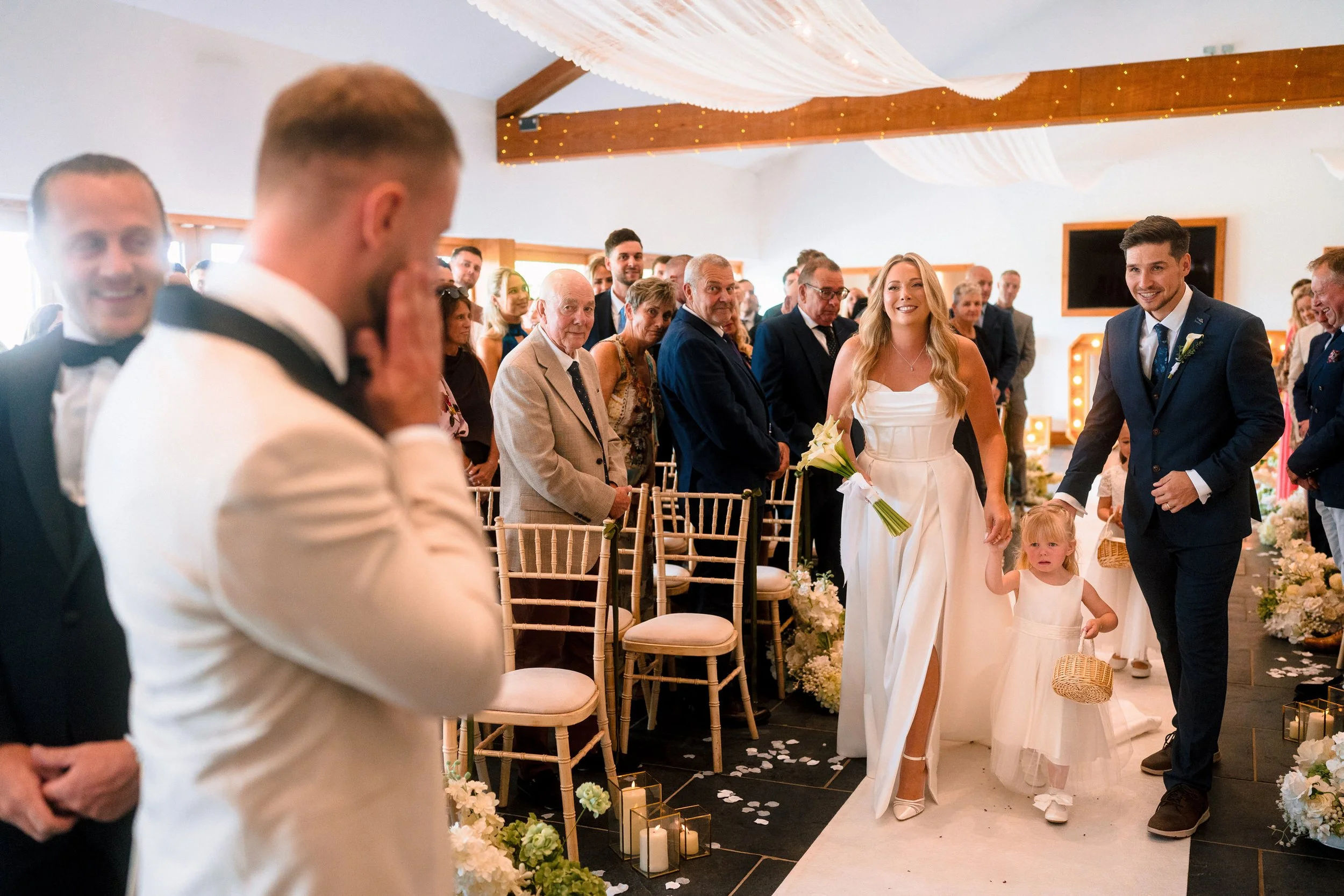 Bride and groom walk down the aisle with a flower girl during an indoor wedding ceremony surrounded by smiling guests.