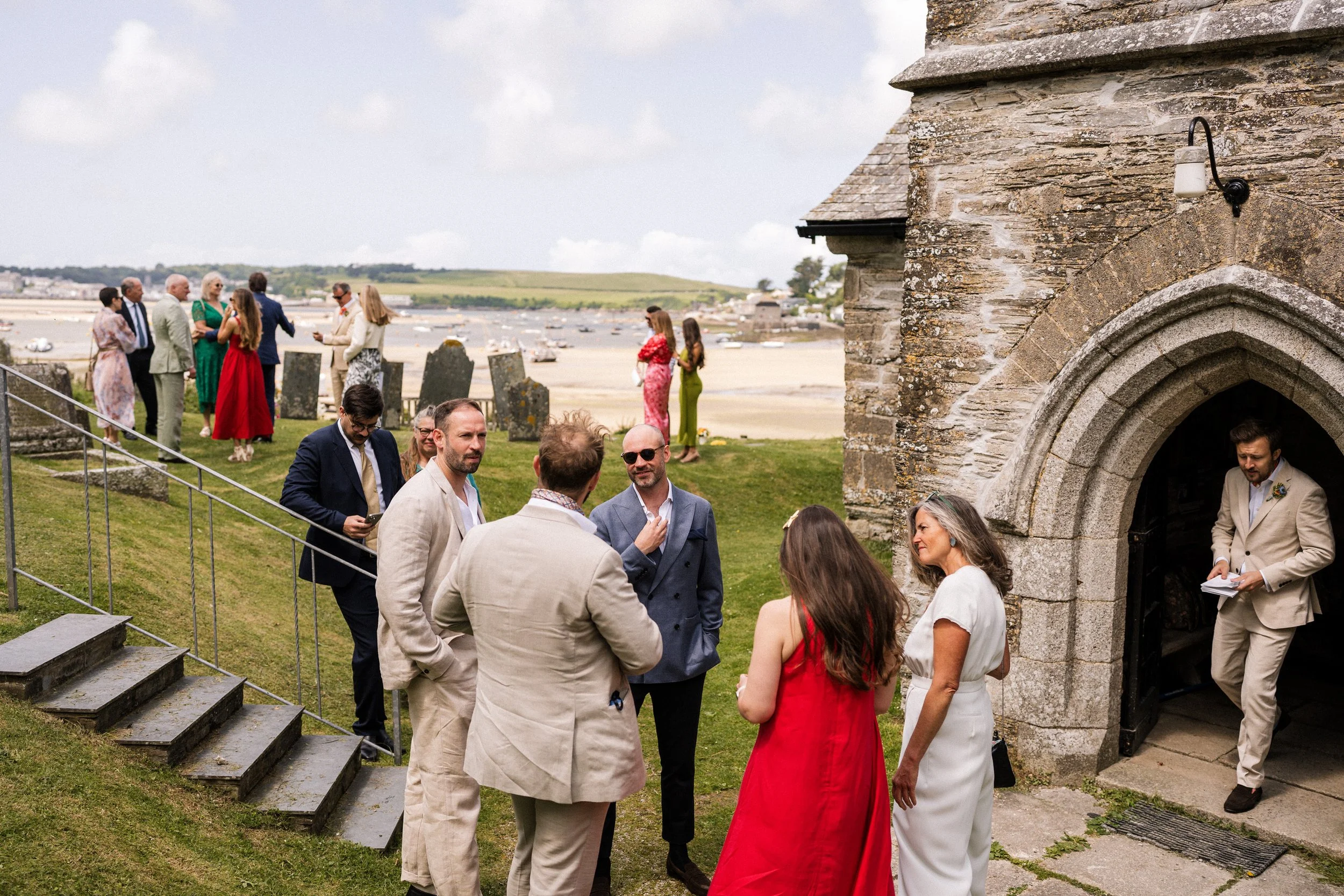 Wedding guests gather outside a historic stone church overlooking a coastal harbor.