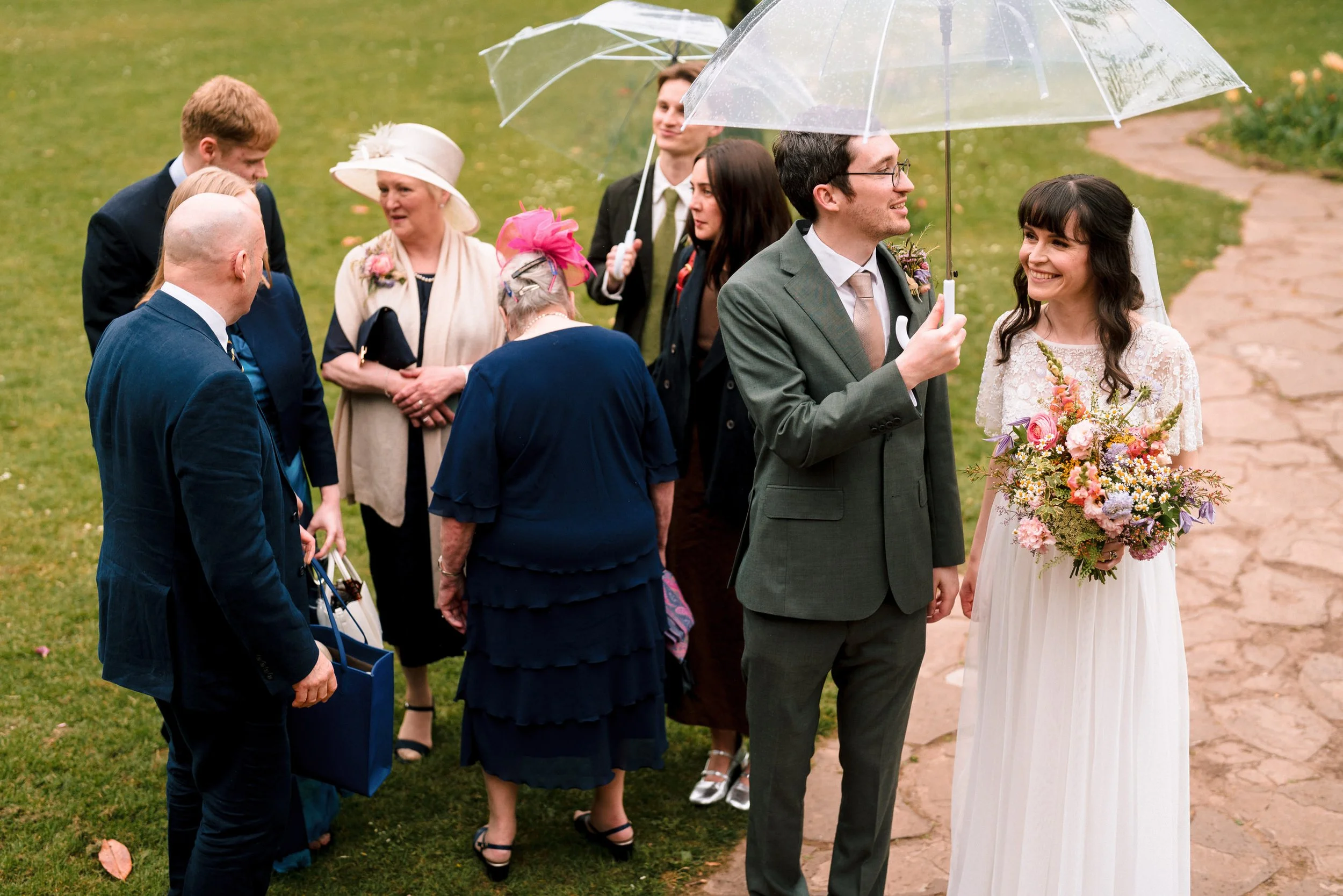 Bride and groom with wedding guests holding clear umbrellas outdoors in a garden.